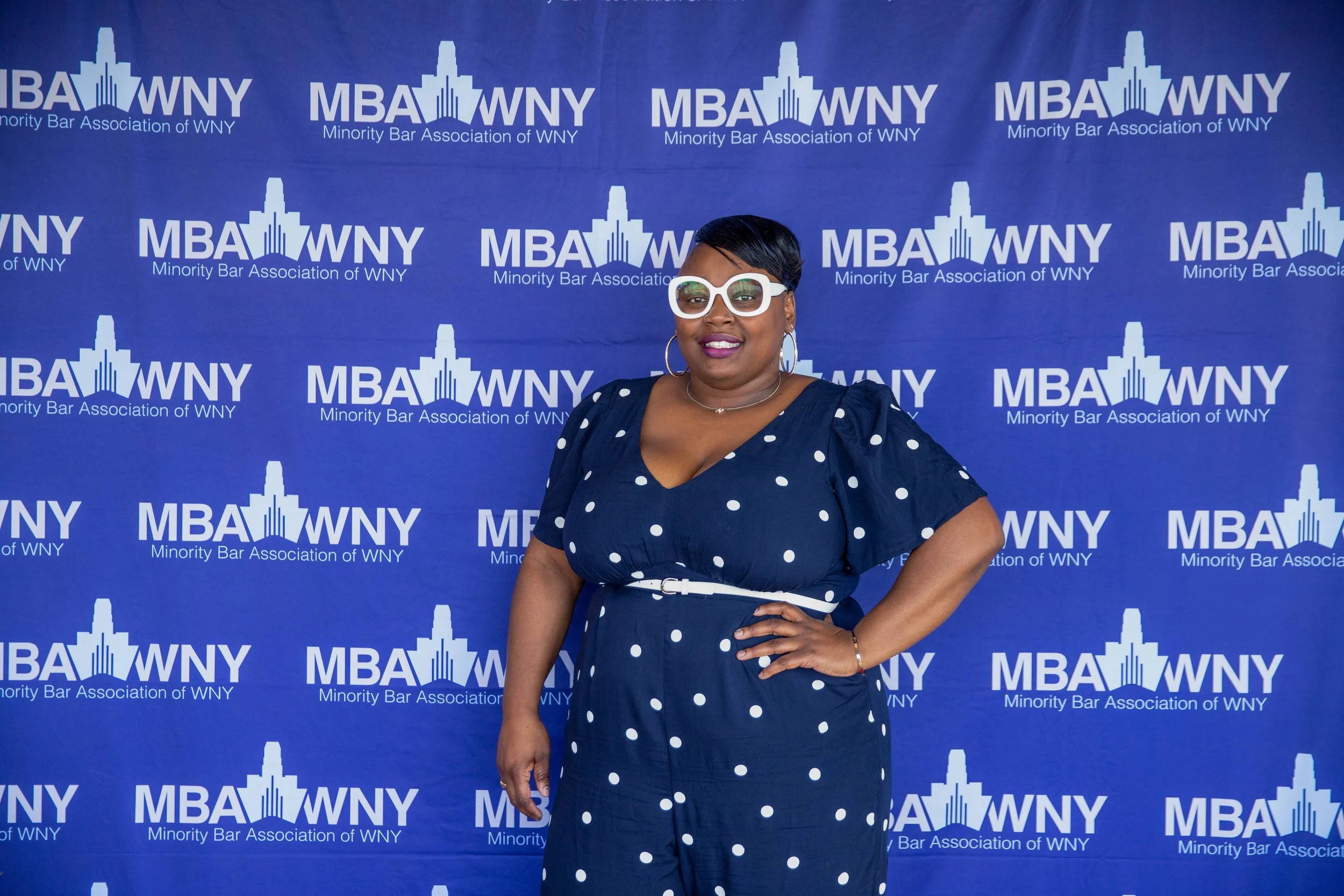 A woman standing confidently in front of a blue backdrop with the logo and text for the Minority Bar Association of WNY. She is smiling, wearing large white glasses, earrings, a navy polka-dot dress with a white belt, and has her hand on her hip.