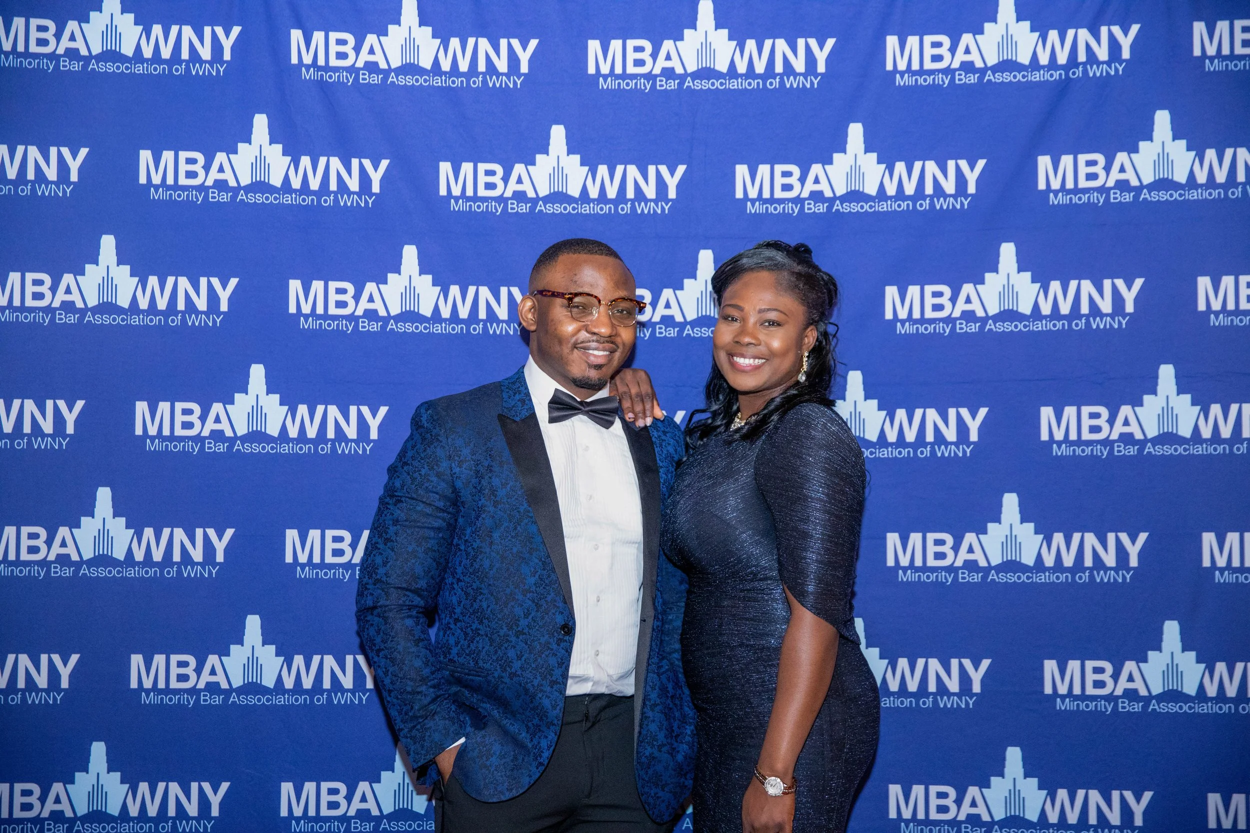 A man and woman dressed formally at an event, standing in front of a blue backdrop with the logo and name of the Minority Bar Association of WNY.