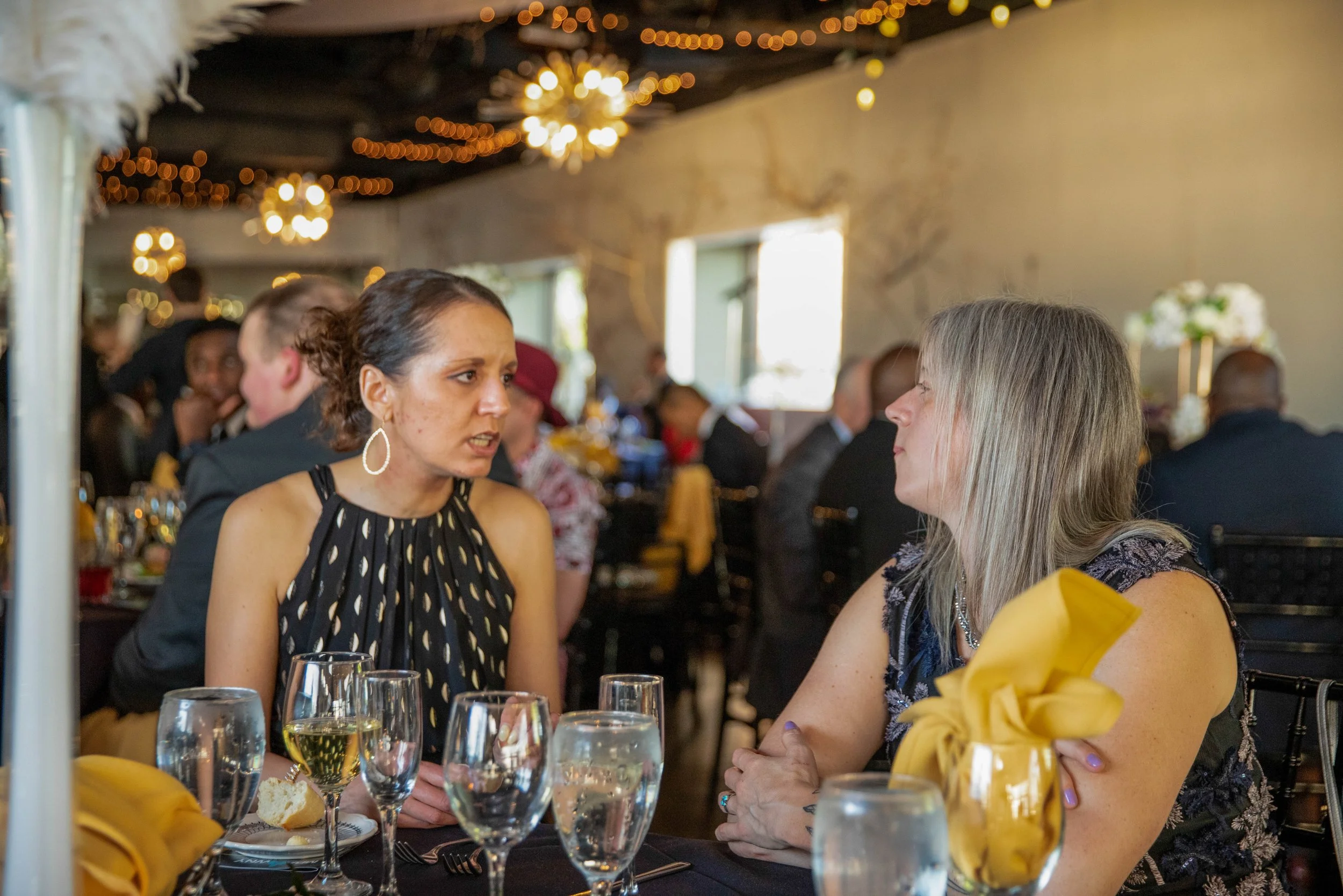 Two women are seated at a table in a decorated dining area, engaging in conversation, with glasses of wine and a plate of bread on the table, while other guests are visible in the background.