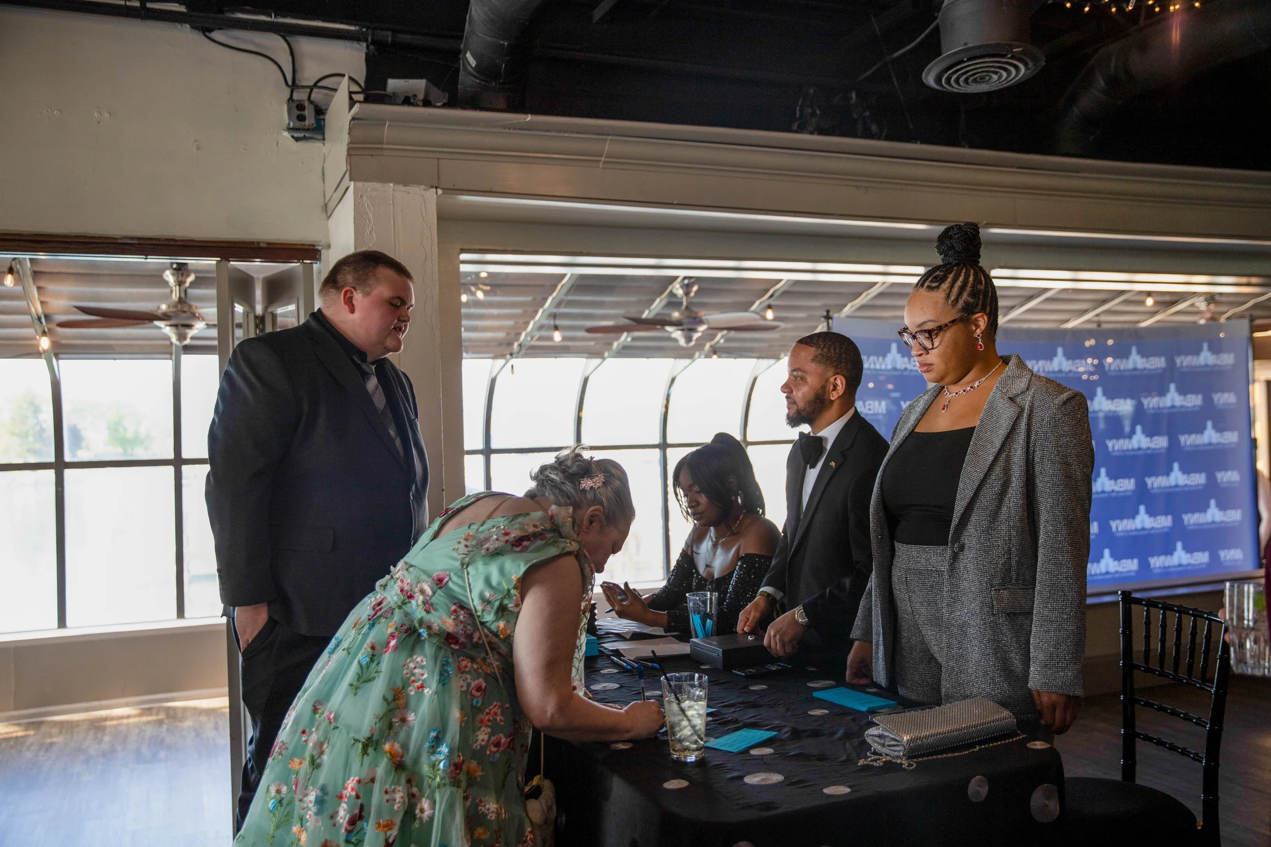People at a registration table during a formal event in a well-lit room with large windows, decorated with a blue background banner.