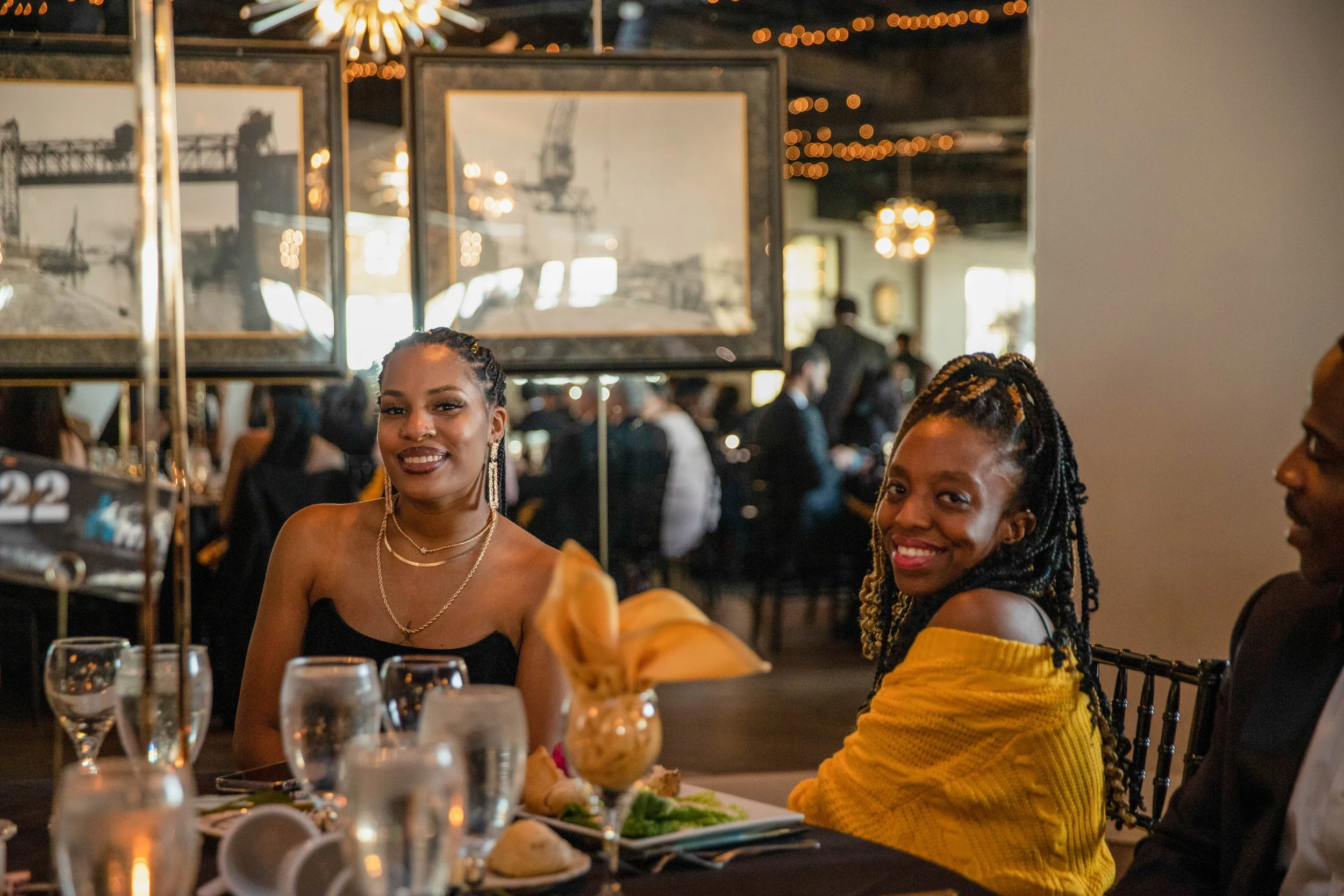 Two women are sitting at a dinner table in a restaurant, smiling at the camera. The woman on the left is wearing a black top with gold jewelry, and the woman on the right is wearing a yellow top with dreadlocks. The table has wine glasses, plates, an