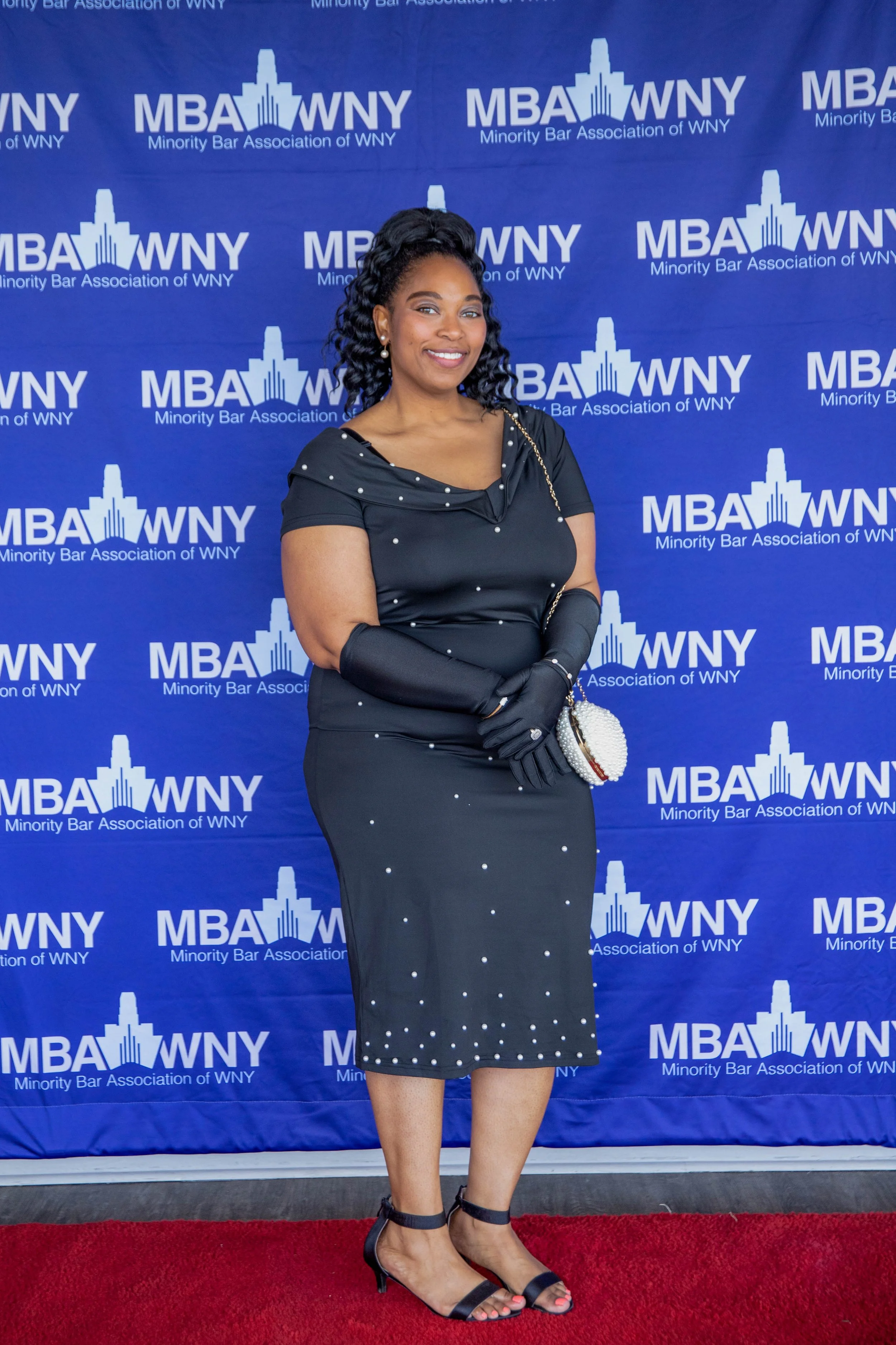 A woman in a black dress with pearl accents and black gloves standing on a red carpet at a formal event. She is holding a pearl clutch purse and wearing black heels. Behind her, a blue backdrop displays the logo for the Minority Bar Association of WN