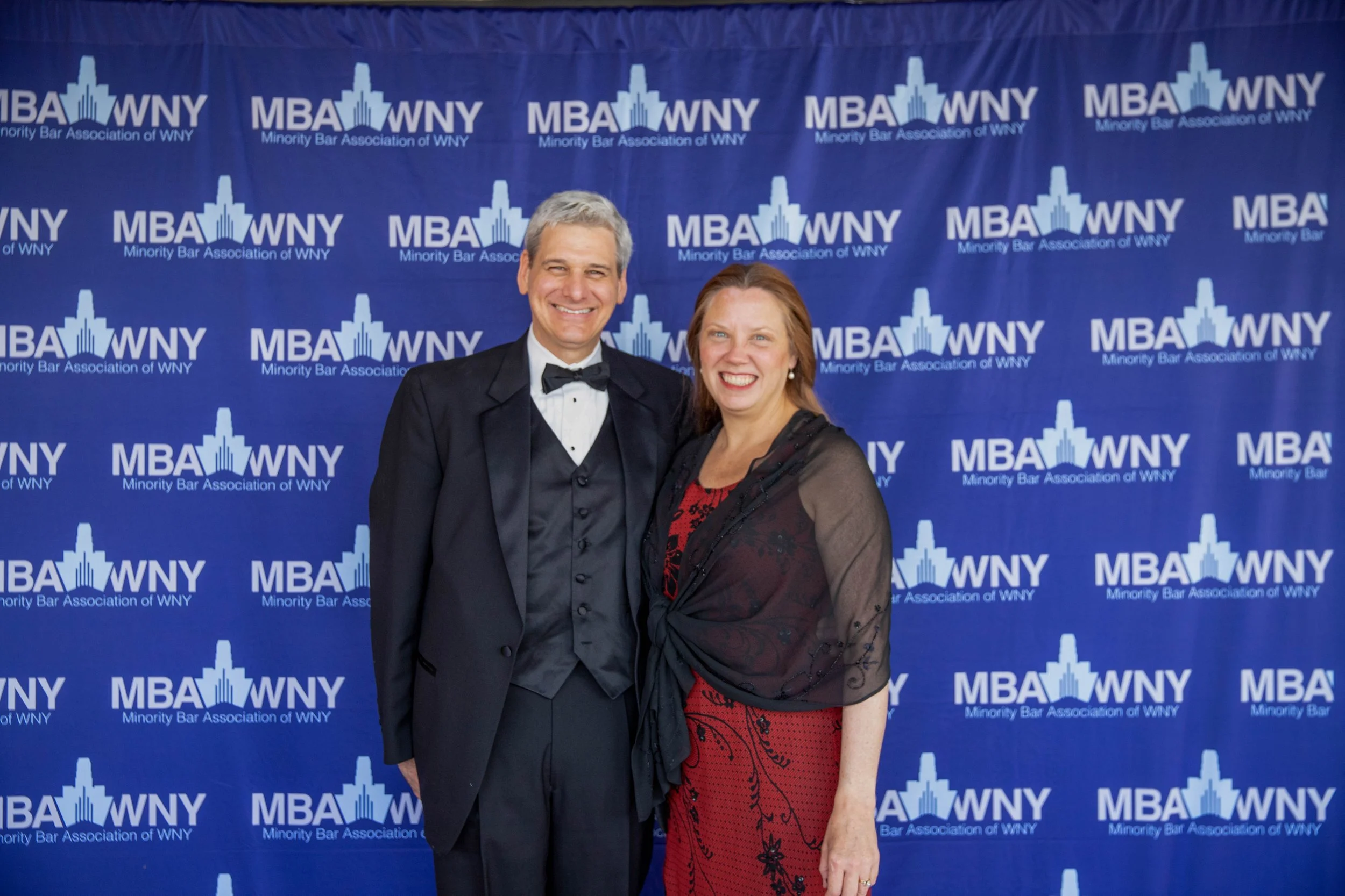 A man in a tuxedo and a woman in a red dress with black lace standing together, smiling at the camera at an event for the Minority Bar Association of WNY, with a blue backdrop featuring their logo.