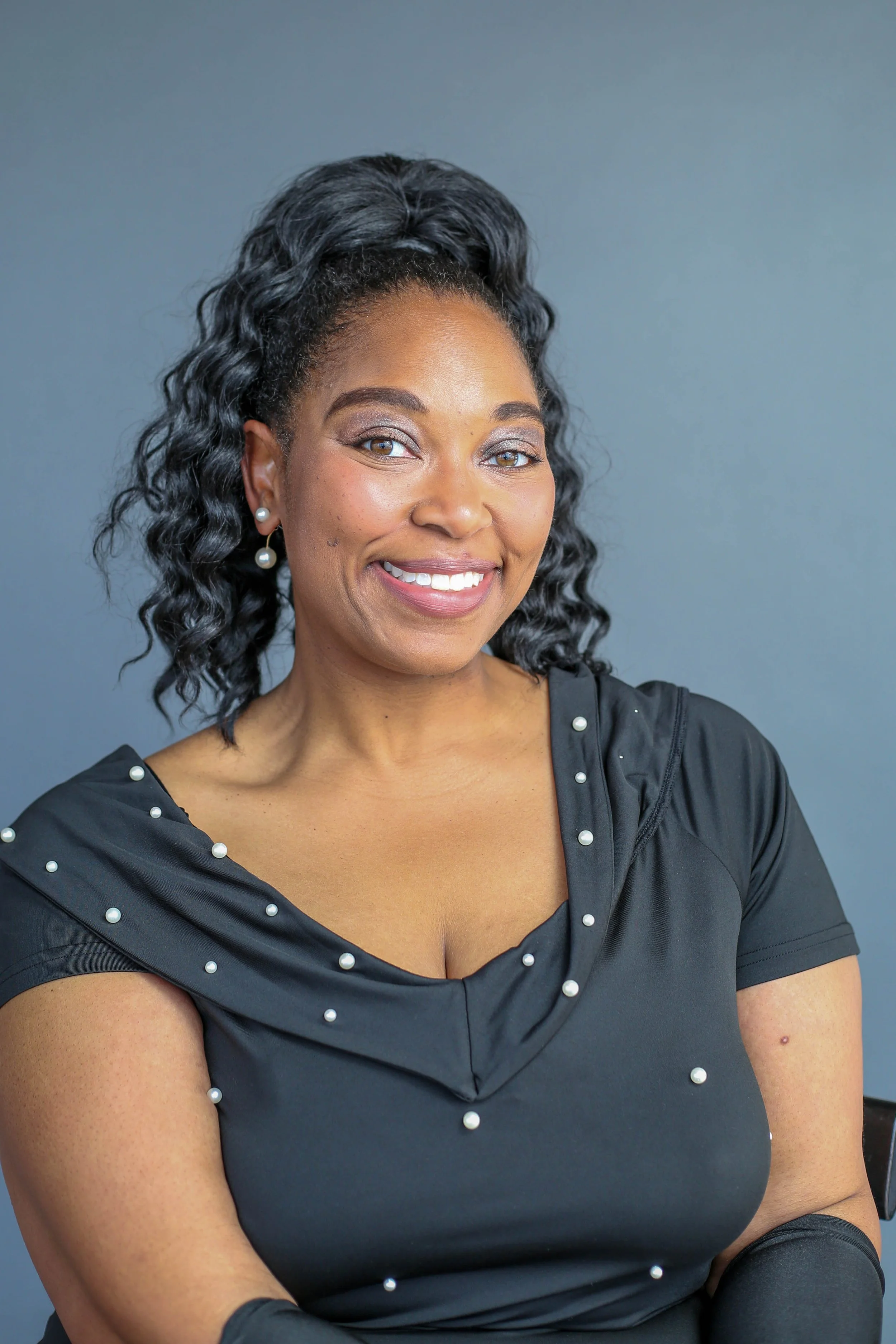 A smiling woman with medium brown skin, curly black hair tied up in a high ponytail, wearing pearl earrings and a black dress decorated with pearl accents, standing against a plain grey background.
