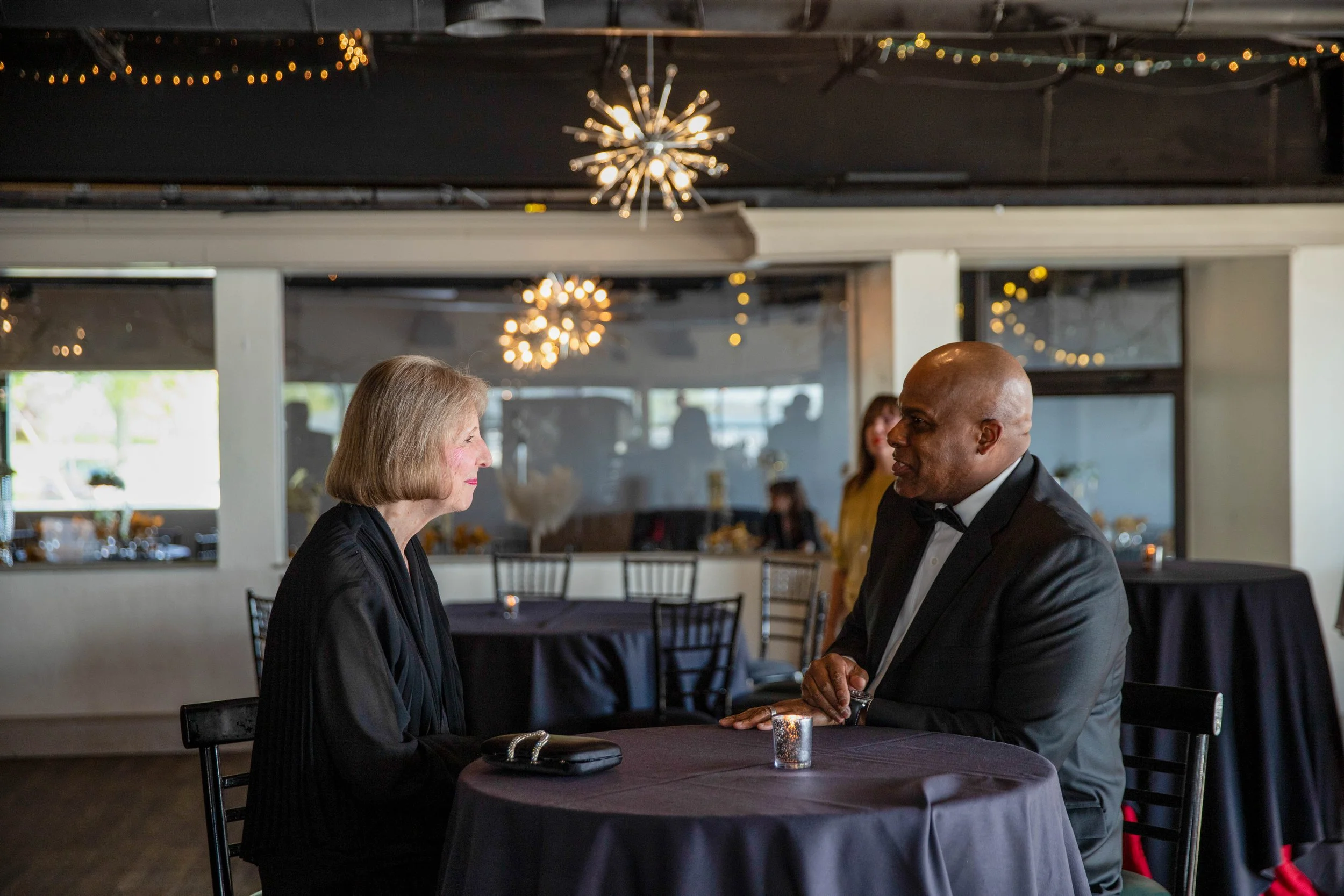An elderly woman and a middle-aged man are sitting across from each other at a restaurant table, holding hands and gazing at each other. The man is wearing a tuxedo, and the woman is dressed in a black outfit. The restaurant has modern decor with str
