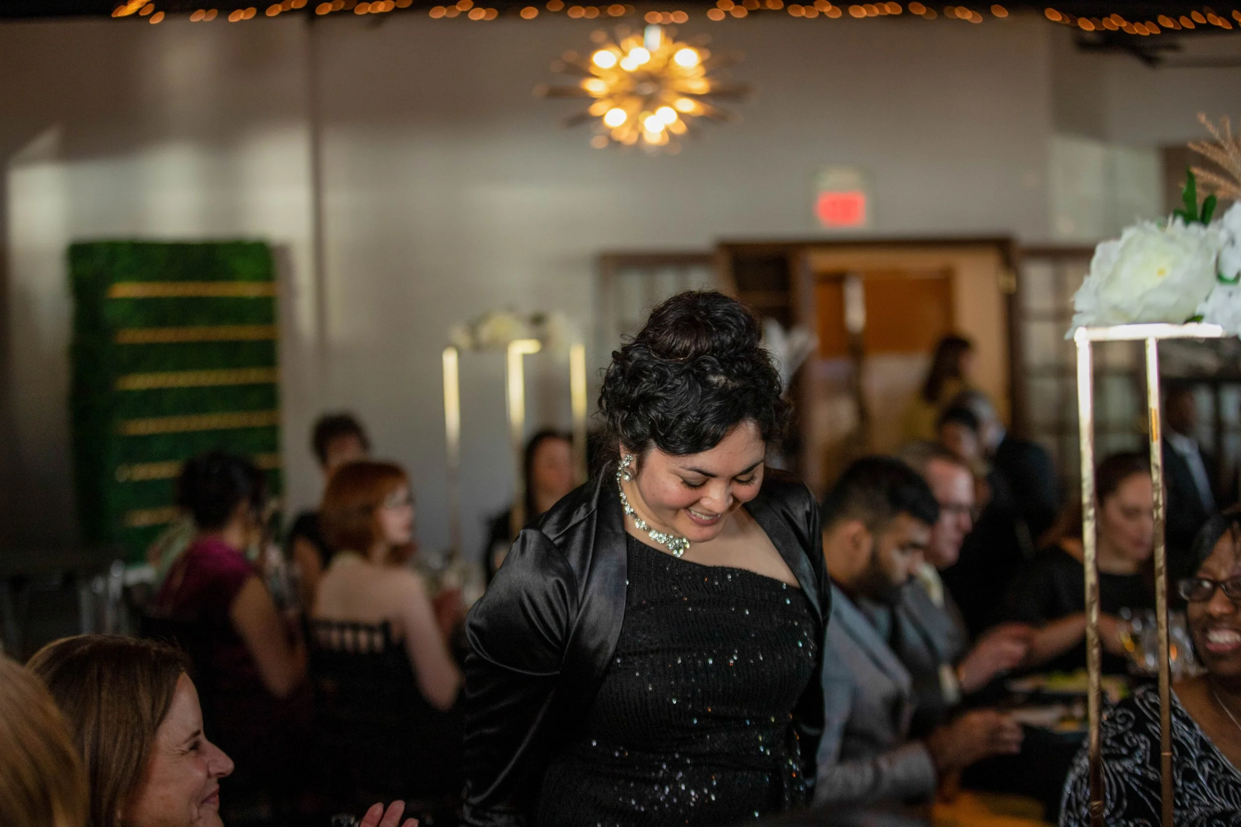 A woman with short dark hair smiling, dressed in a black dress with a shiny necklace, attending a crowded indoor event with many people seated, decorative elements, and warm lighting.