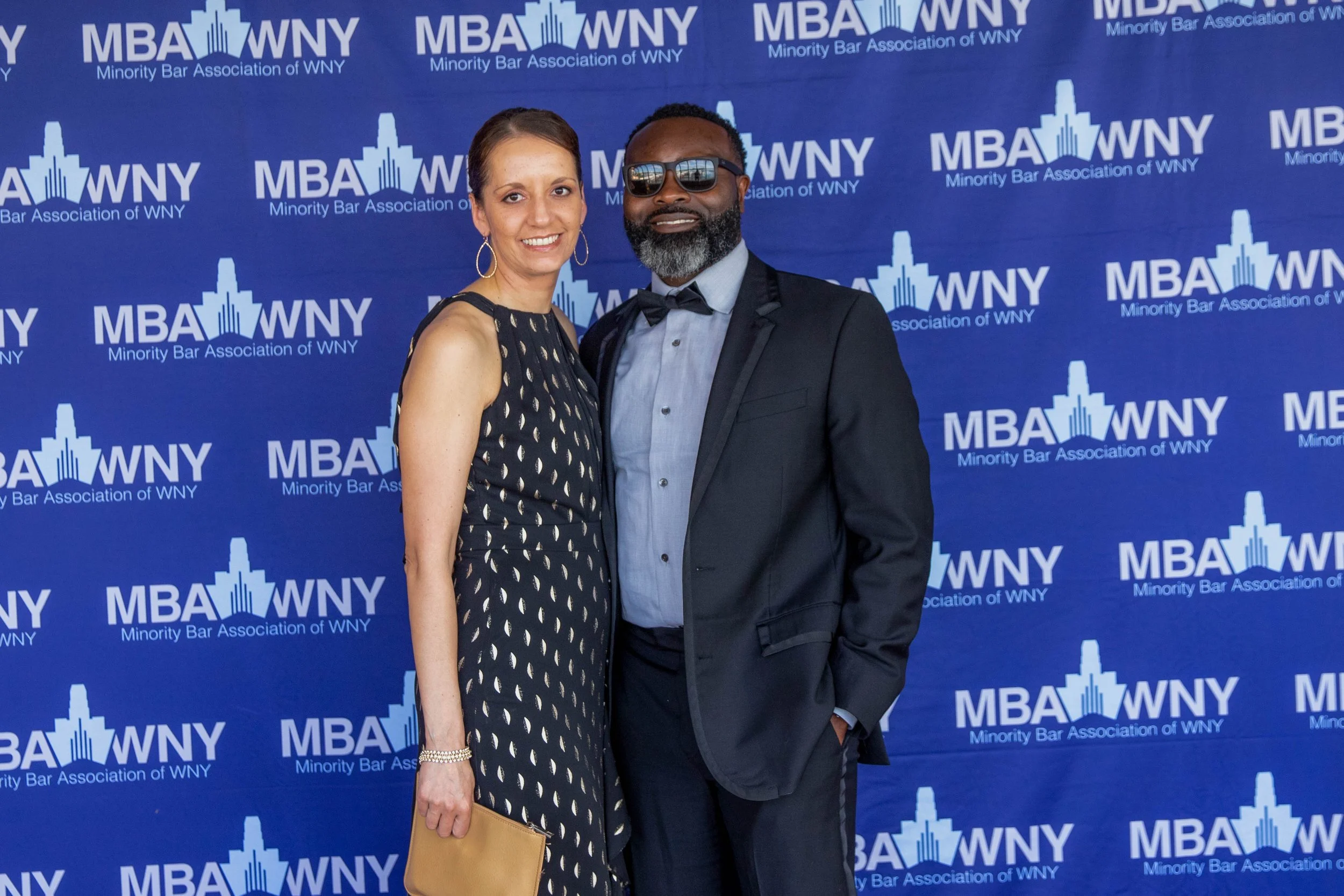 A man and woman dressed in formal attire standing in front of a blue backdrop with the MBA WNY logo and text for the Minority Bar Association of WNY.