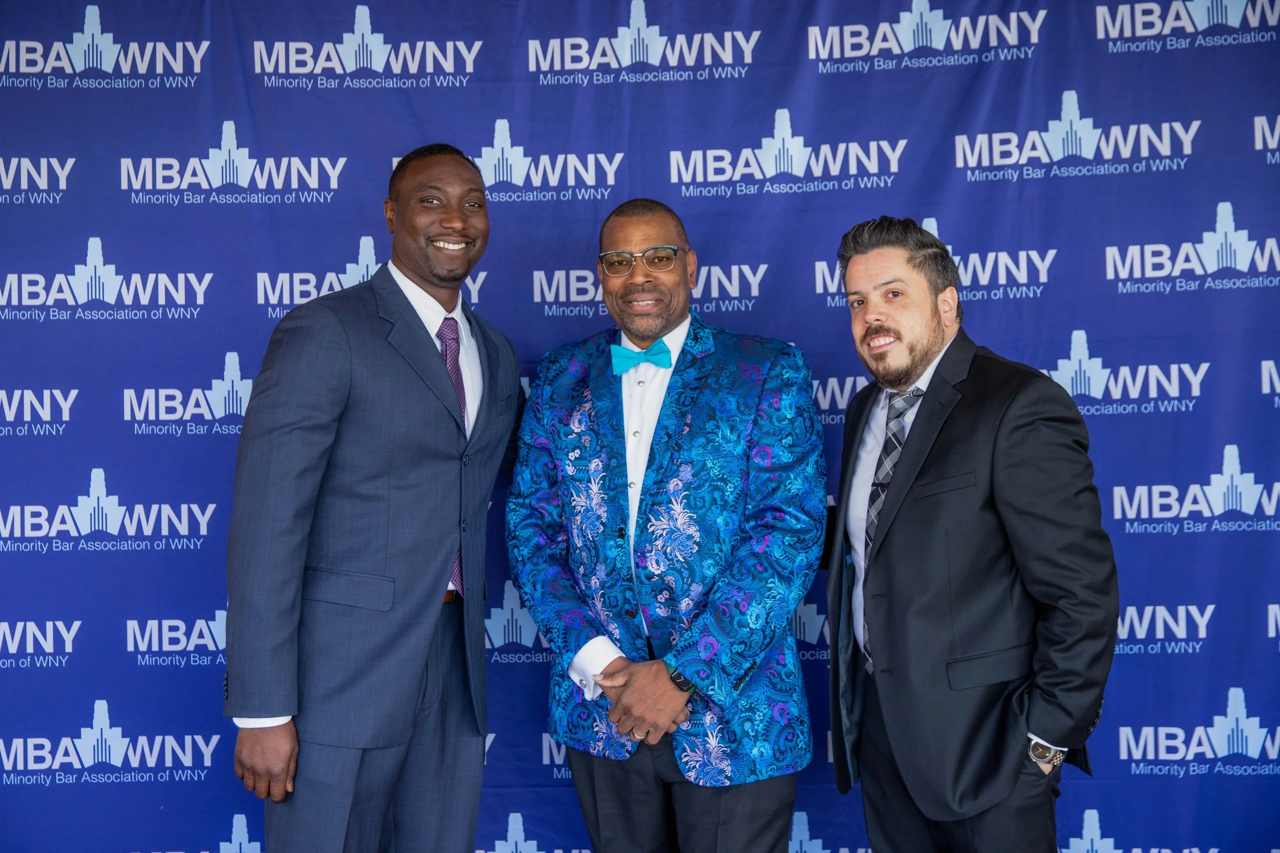 Three men in suits standing in front of a blue backdrop with the logo and text for the Minority Bar Association of WNY, posing for a photo at an event.