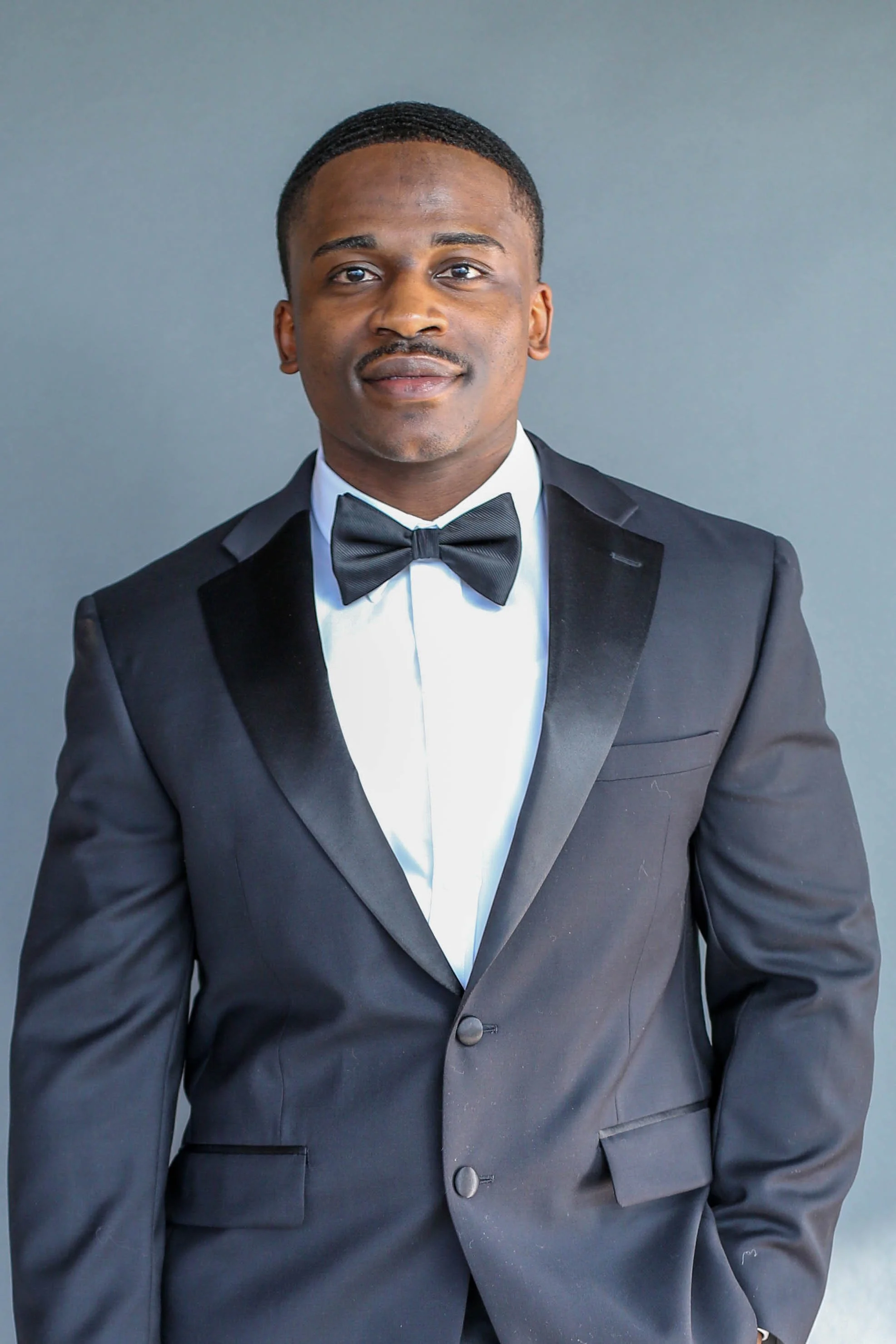 A young man wearing a tuxedo with a bow tie, against a neutral background.