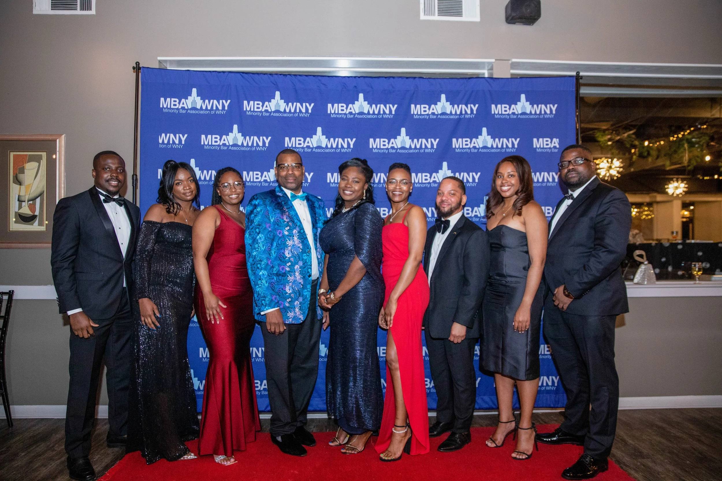 Group of nine individuals dressed in formal attire standing on a red carpet in front of a blue backdrop with logos that read 'MBA WNY Minority Bar Association of WNY.' They are smiling and posing for a photo indoors.