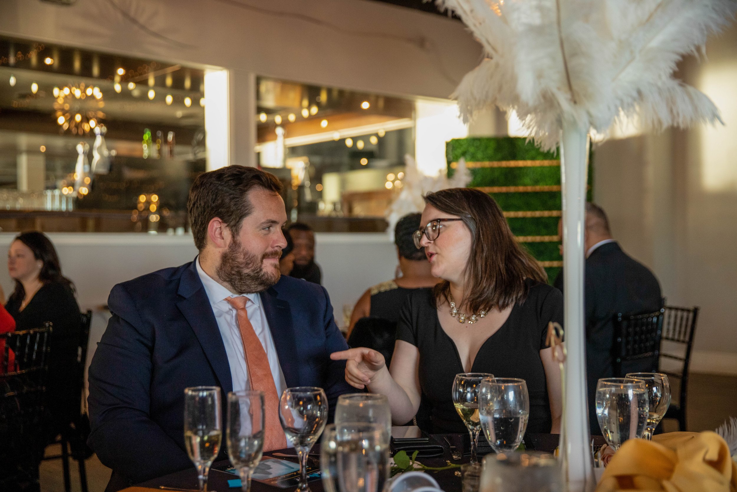 A man and a woman seated at a table in a restaurant, engaging in conversation. The table has several glasses of water, a yellow tablecloth, and a white feathered umbrella. The background shows other guests and warm lighting fixtures in a modern, styl