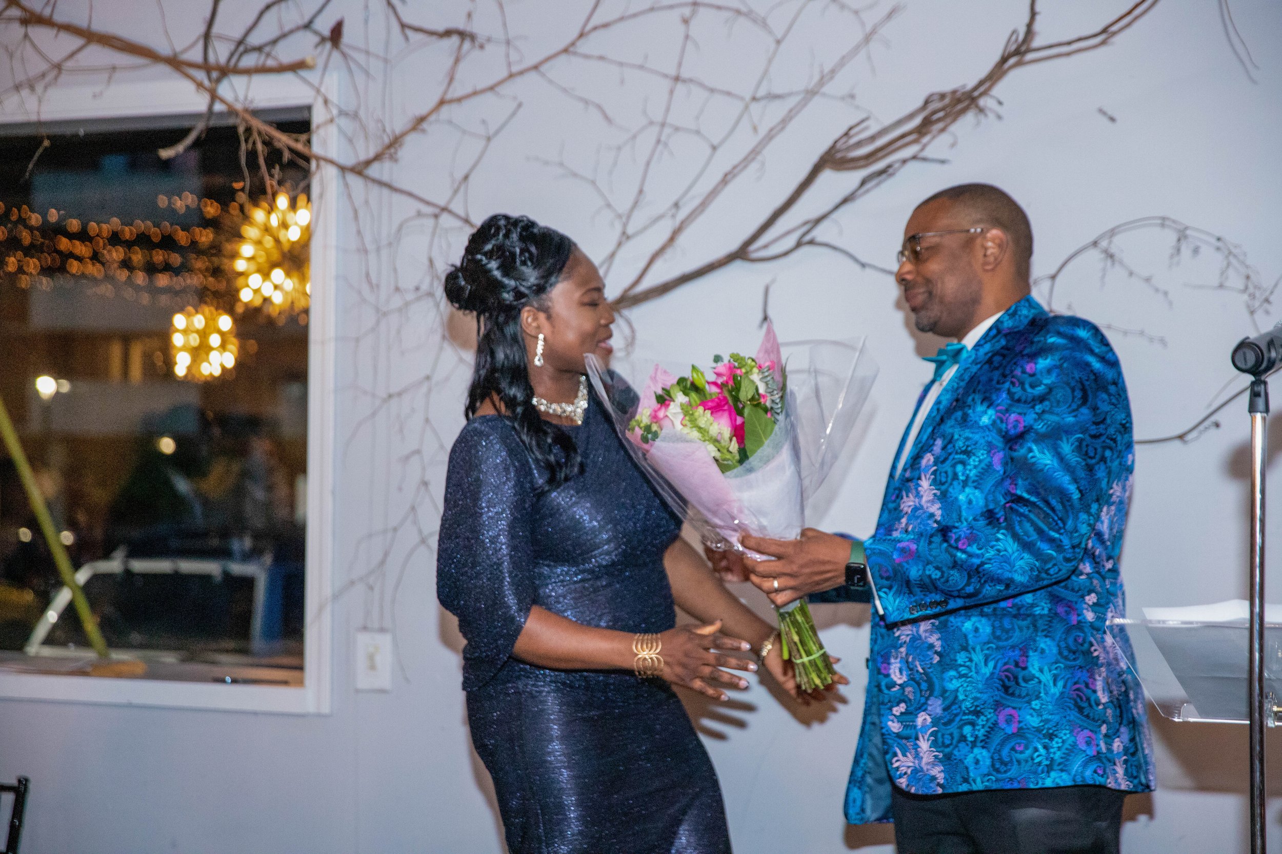 A woman in a dark, shiny dress receiving a bouquet of pink flowers from a man in a blue patterned suit during a celebration or ceremony.