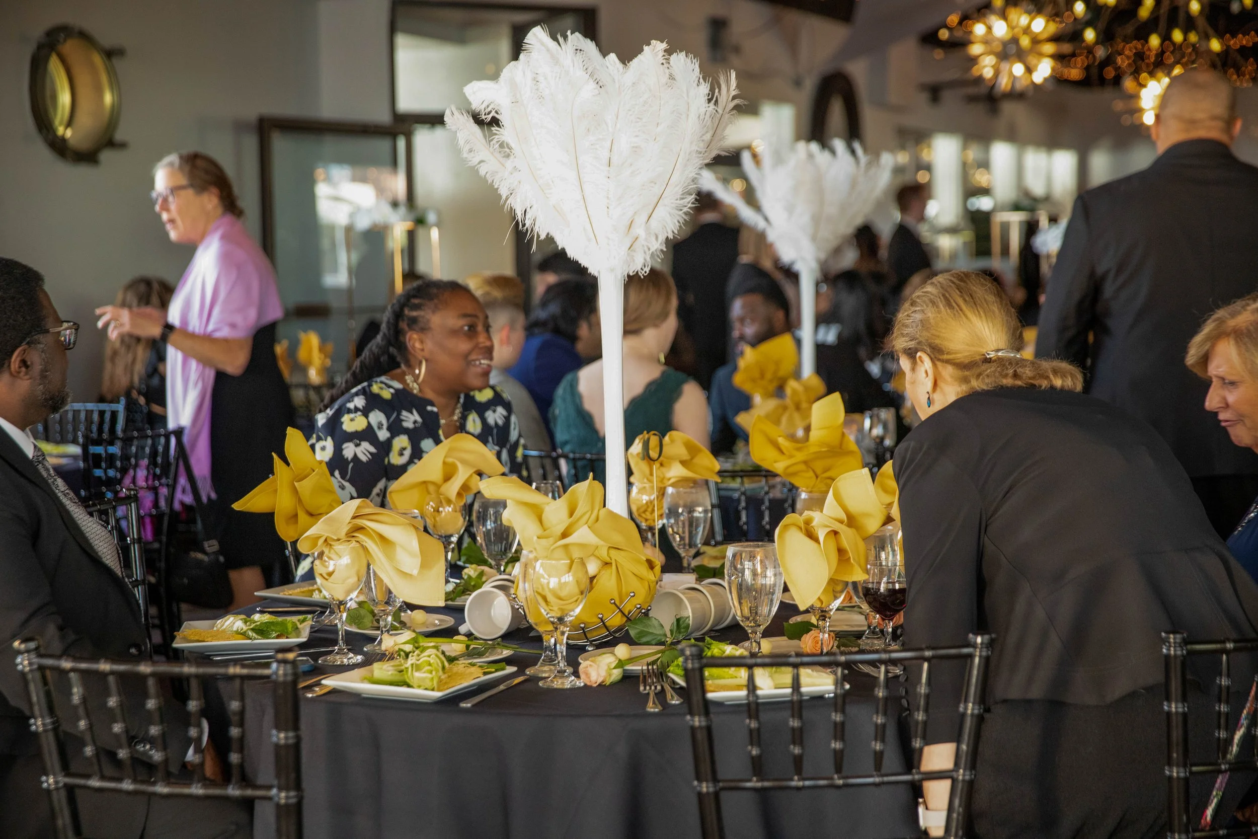 People gathered at a formal event with decorated tables, centerpiece with white feathers, yellow napkins, and elegant place settings, in a well-lit indoor venue.
