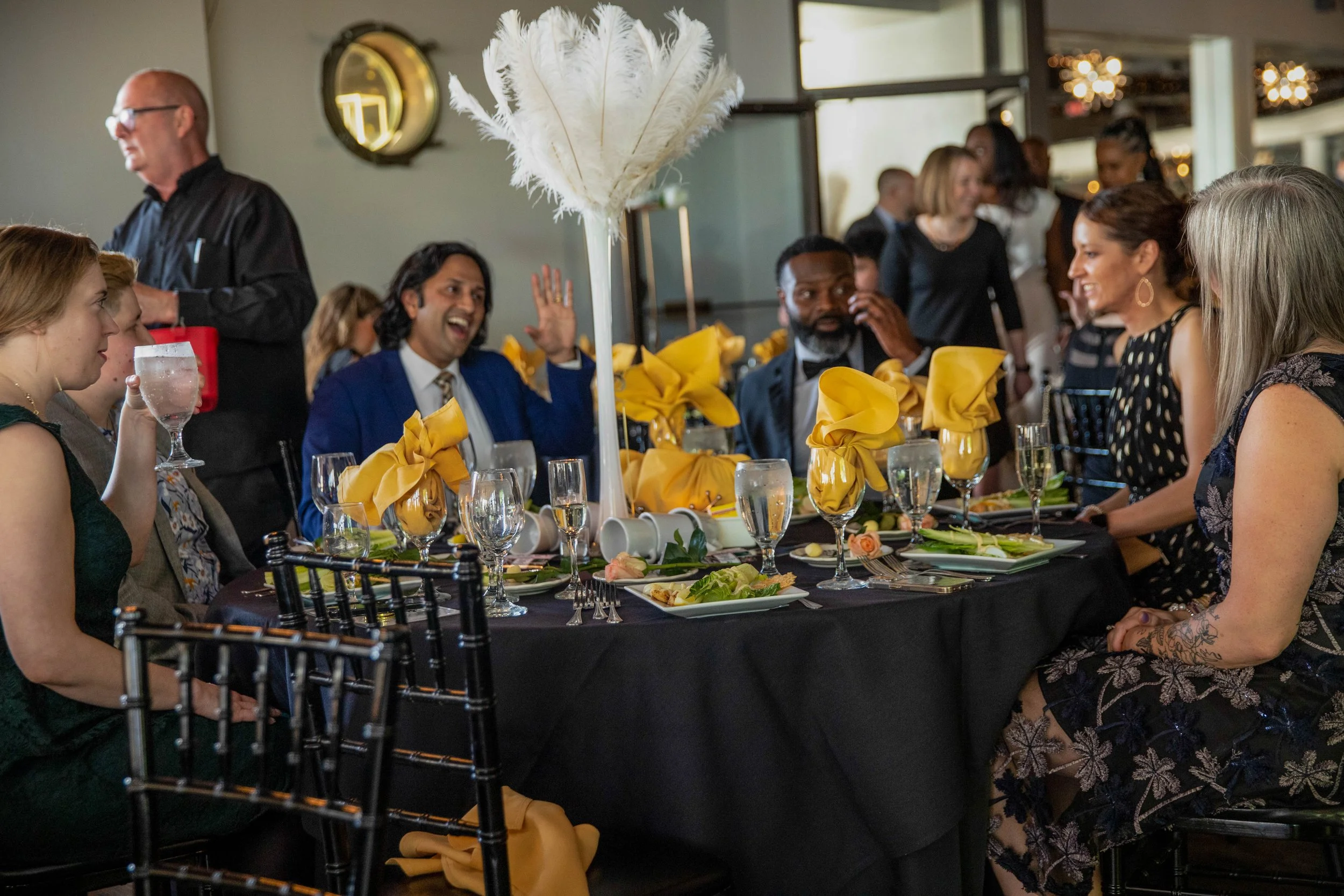 People dining at a dinner event with elegant table settings, floral centerpieces, and guests dressed in formal attire, engaged in conversation and enjoying the occasion.