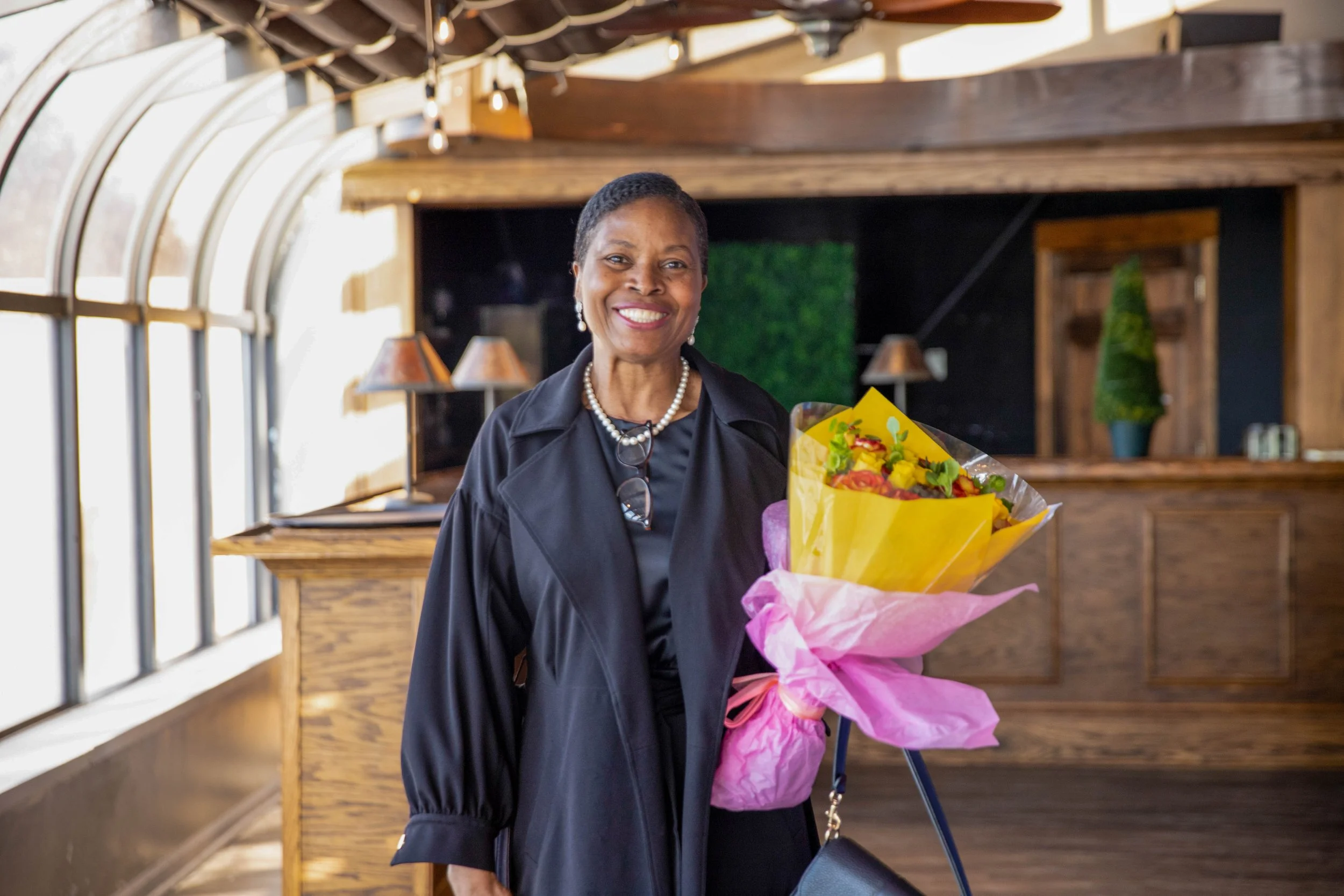 Smiling woman wearing black coat and pearl necklace holding a bouquet of flowers inside a room with wooden walls and large windows.