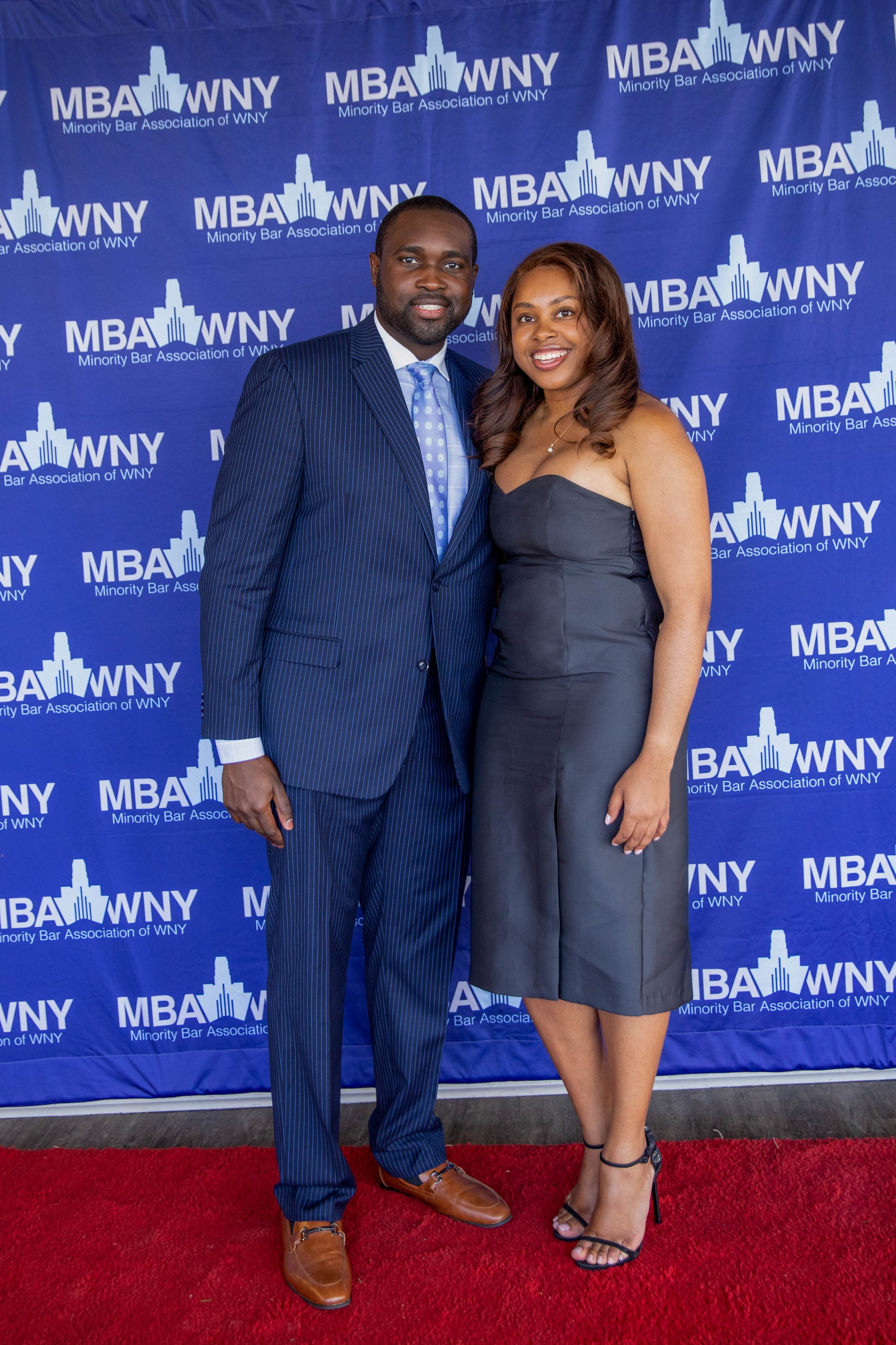 A man and woman are standing on a red carpet in front of a blue backdrop with the logo and text for the Minority Bar Association of WNY. The man is wearing a dark pinstripe suit, light blue shirt, and blue tie. The woman is dressed in a strapless dar
