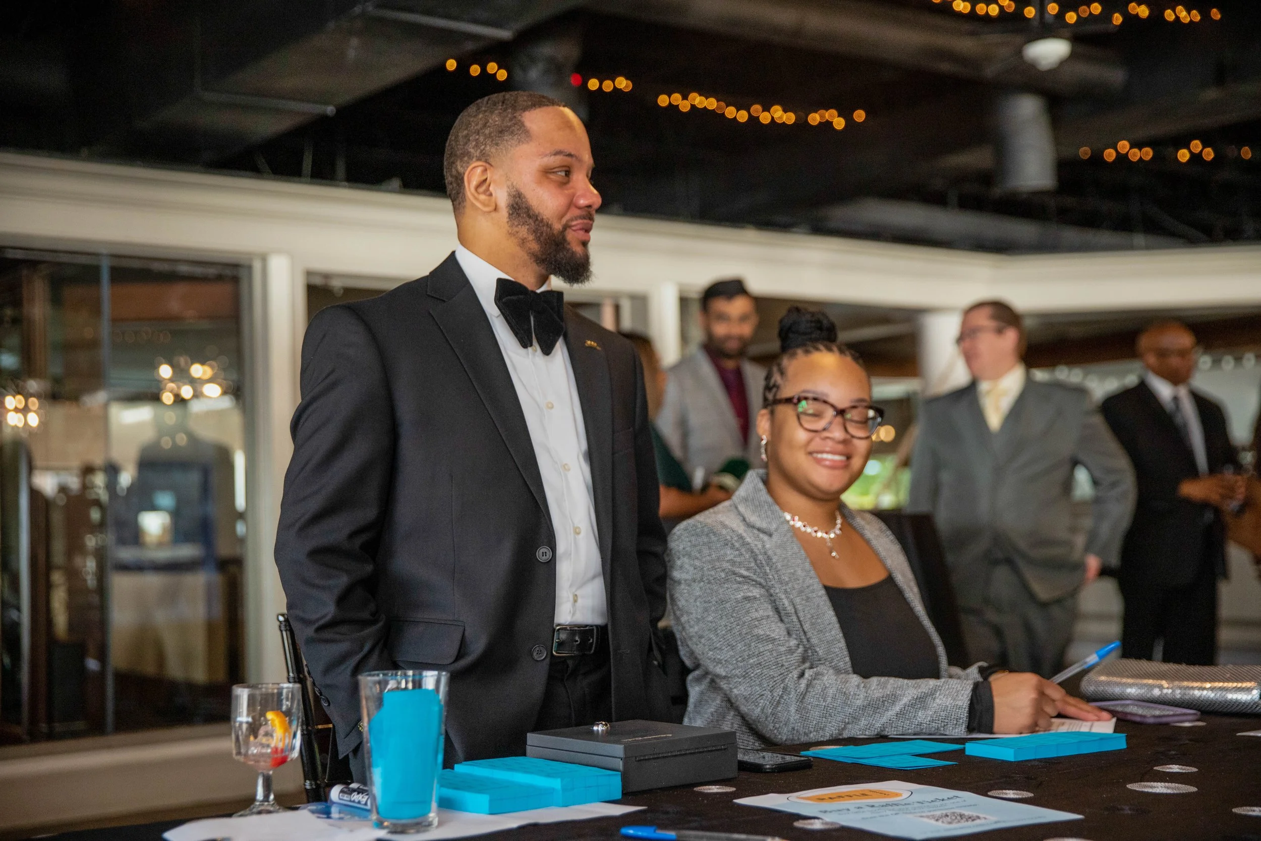 A man in a black tuxedo and bow tie and a woman in glasses and a gray blazer are standing at a table during an event, smiling. Other people are in the background, and the setting appears to be a banquet or formal gathering space with string lights on