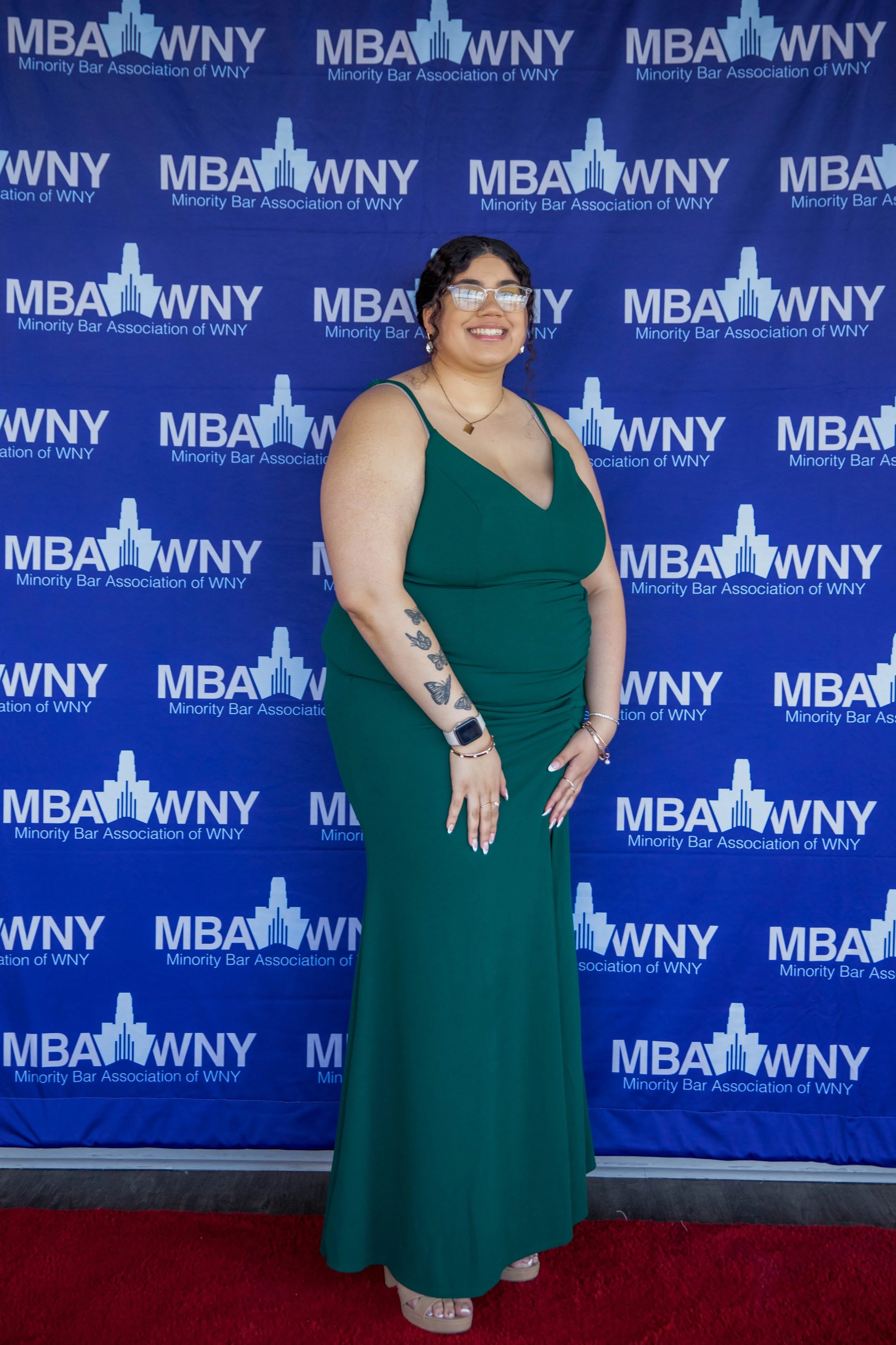 A woman in a green dress stands on a red carpet in front of a blue backdrop with the logos of the Minority Bar Association of WNY. She is smiling, wearing glasses, jewelry, and has visible tattoos on her left arm.