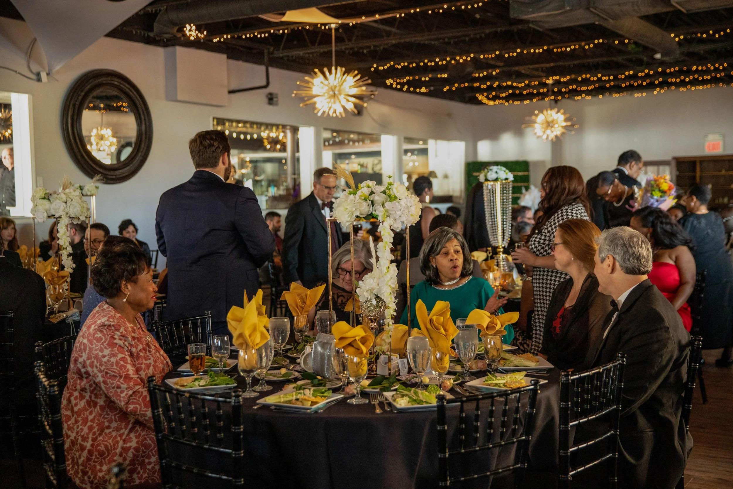 A lively indoor celebration with diverse guests seated at tables decorated with yellow napkins, white floral centerpieces, and plates of food. The room features string lights and modern chandeliers, with some guests standing and socializing.