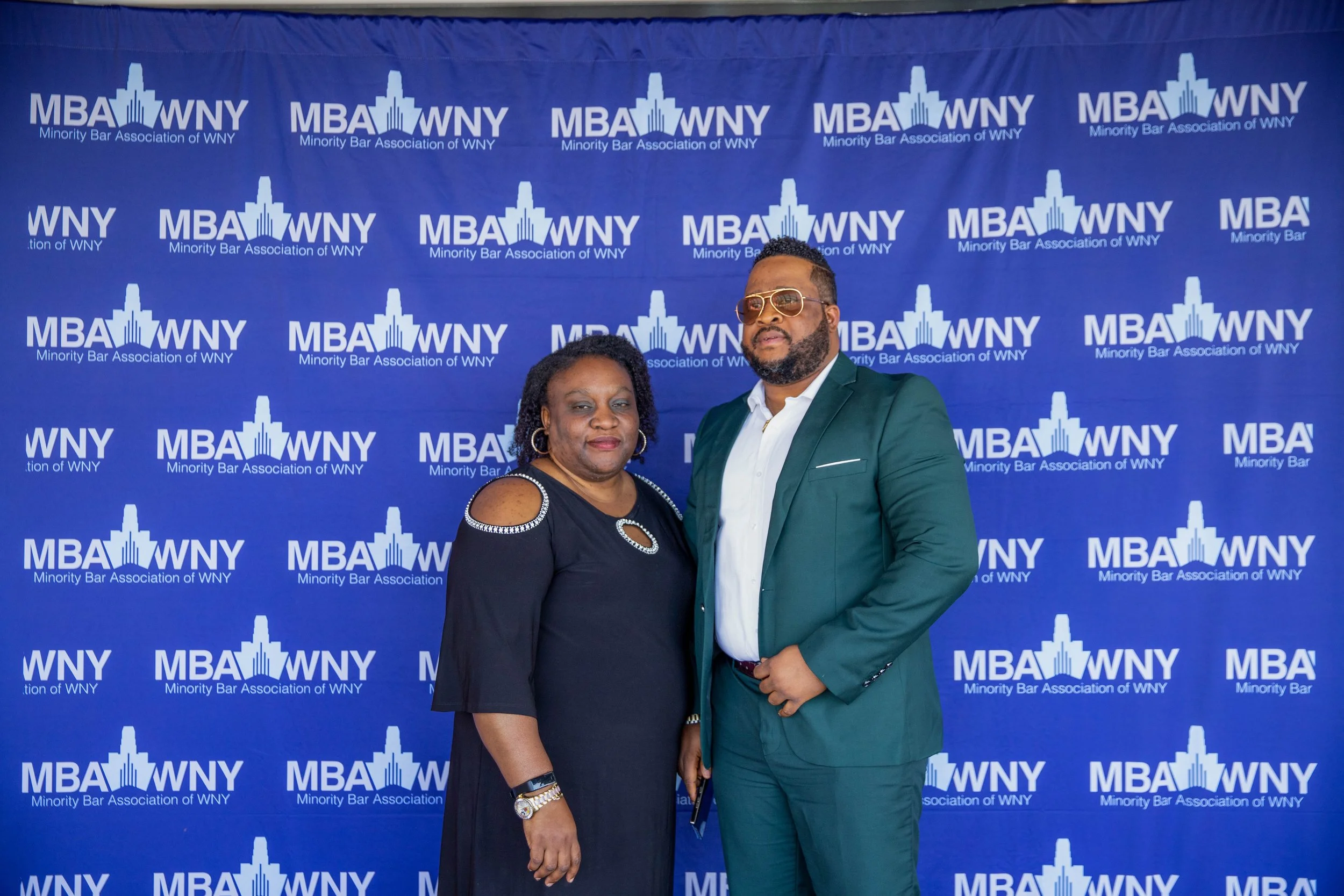 Two people standing in front of a blue backdrop with the logo and text of MBA WNY, representing the Minority Bar Association of WNY. The woman on the left has dark skin and curly hair, wearing earrings and a black top with decorative cutouts on the s