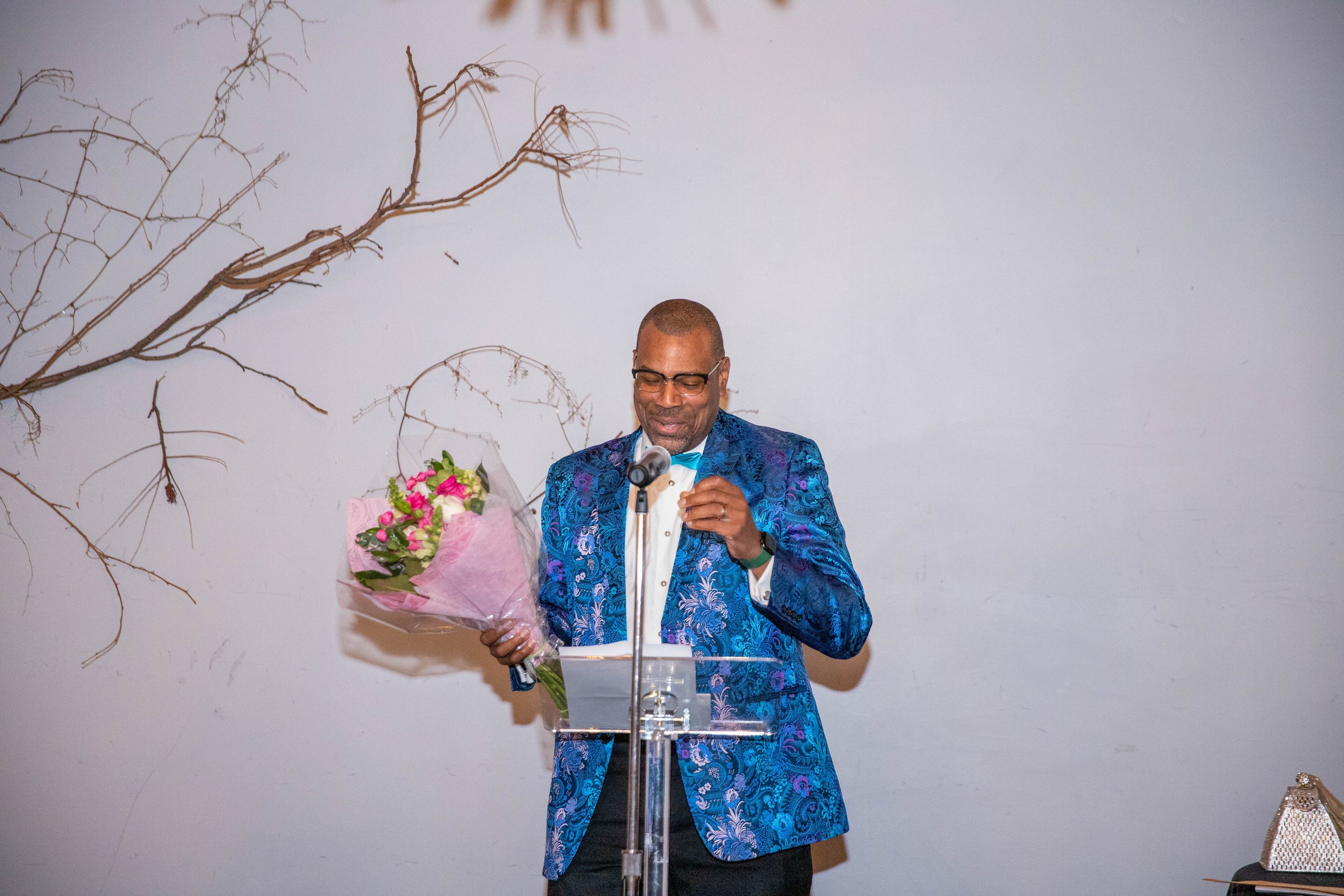 A man in a blue patterned suit and black pants is standing behind a transparent podium, holding a bouquet of pink and white flowers, and speaking into a microphone.
