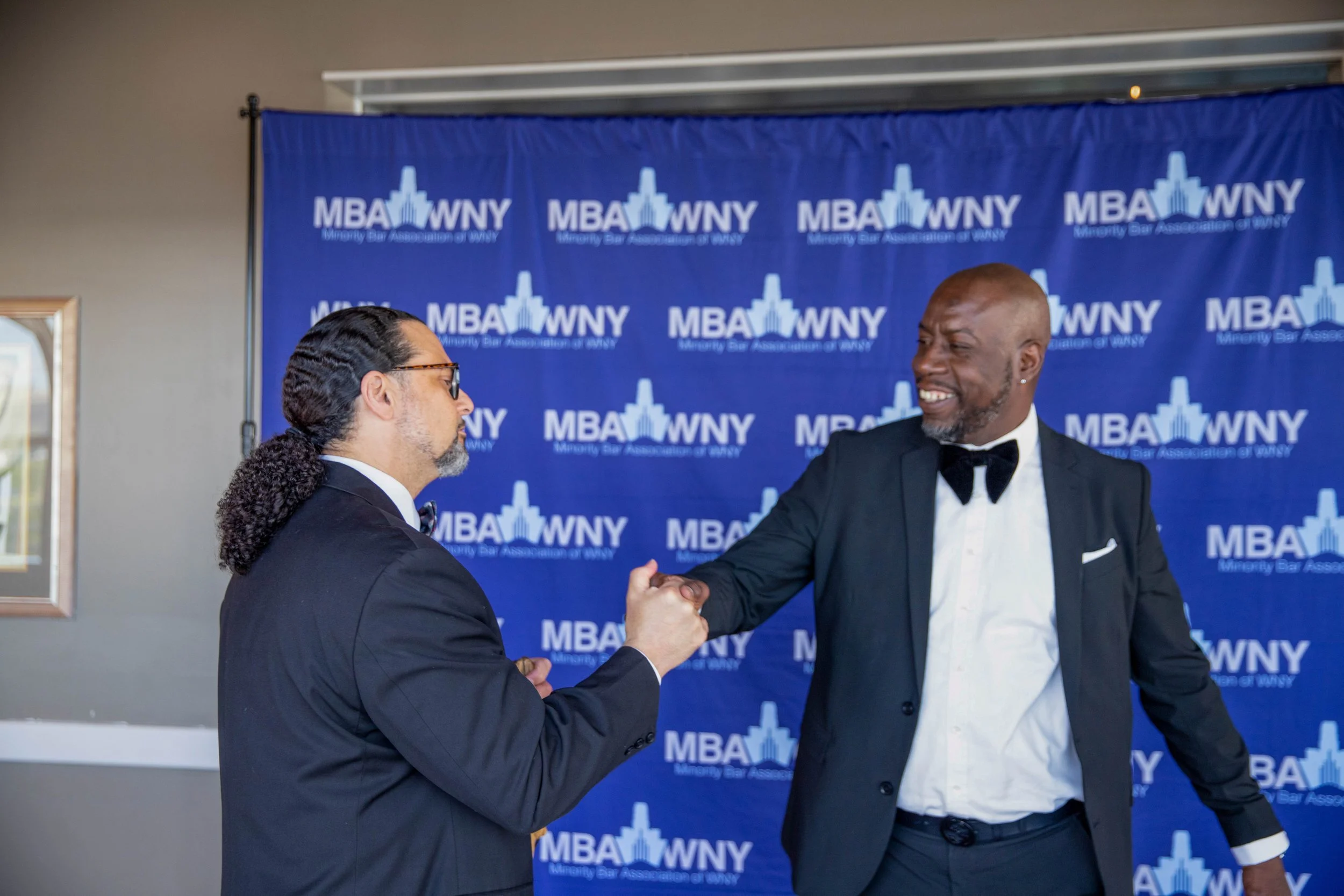 Two men dressed in formal tuxedos shaking hands in front of a blue banner with the text "MBA WNY Minority Bar Association of WNY" and a cityscape logo.