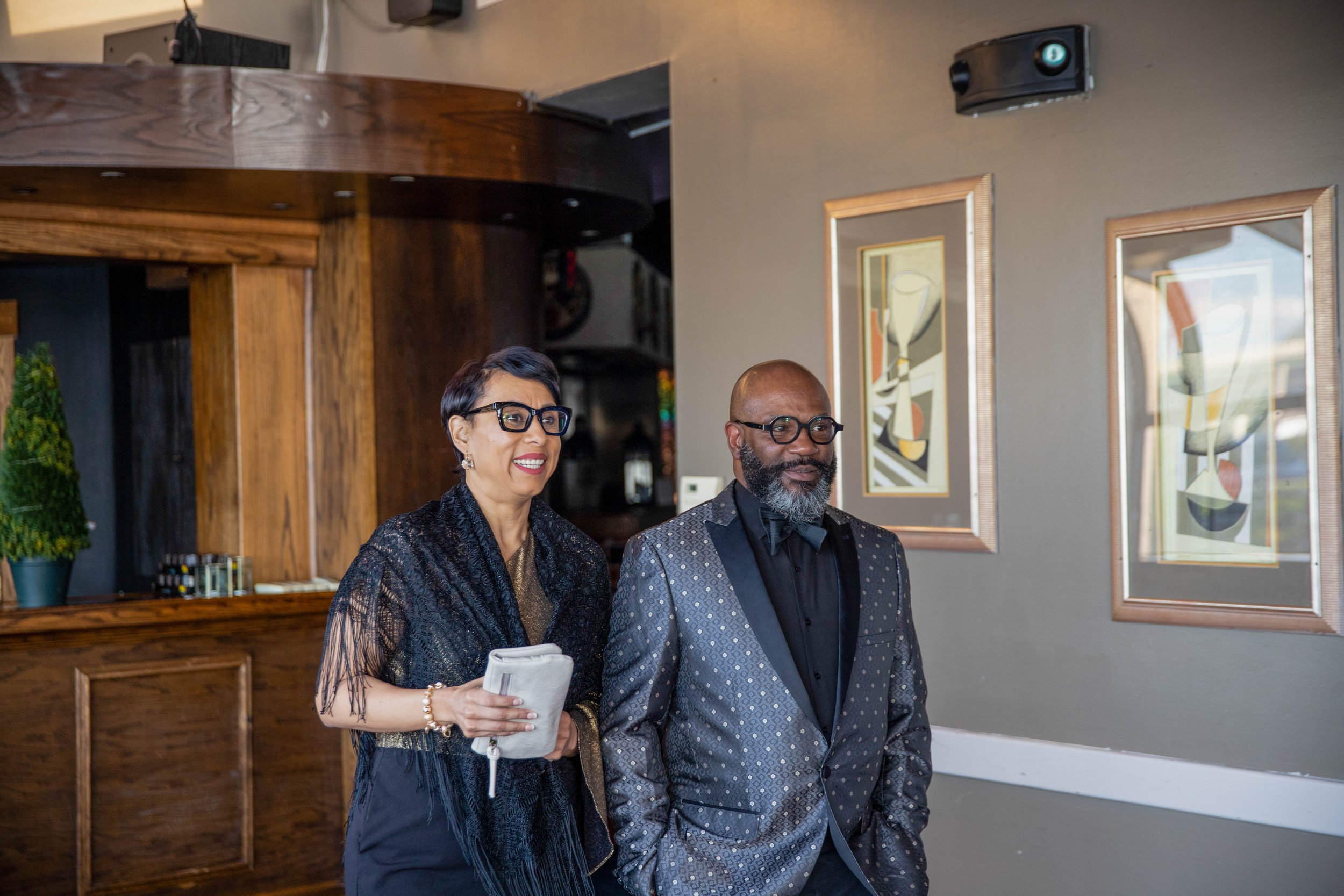 An older woman with short dark hair and glasses, wearing a black dress and pearl jewelry, is holding a white clutch purse and smiling. Next to her is a bald man with a beard, glasses, and a black bow tie, wearing a patterned suit. They are standing i