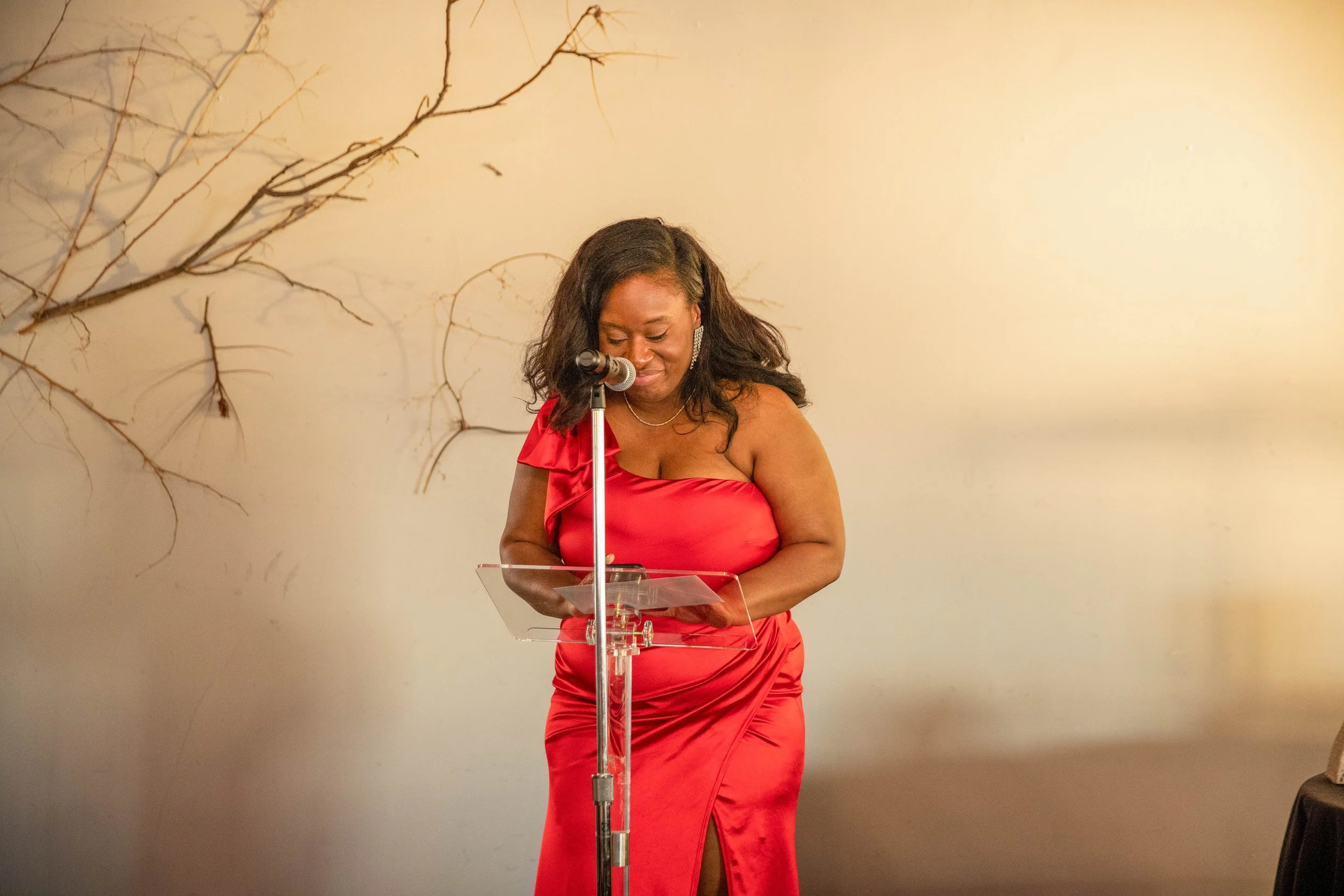 A woman in a red dress standing behind a clear podium, speaking into a microphone, with a decorative branch on the beige wall behind her.