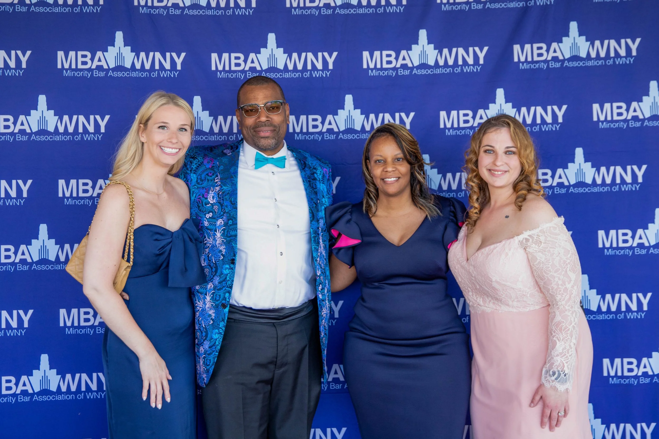 Four people standing in front of a blue background with the text 'Minority Bar Association of WNY' and 'MBA WNY'. They are dressed in formal attire, smiling, with their arms around each other, at a professional event.