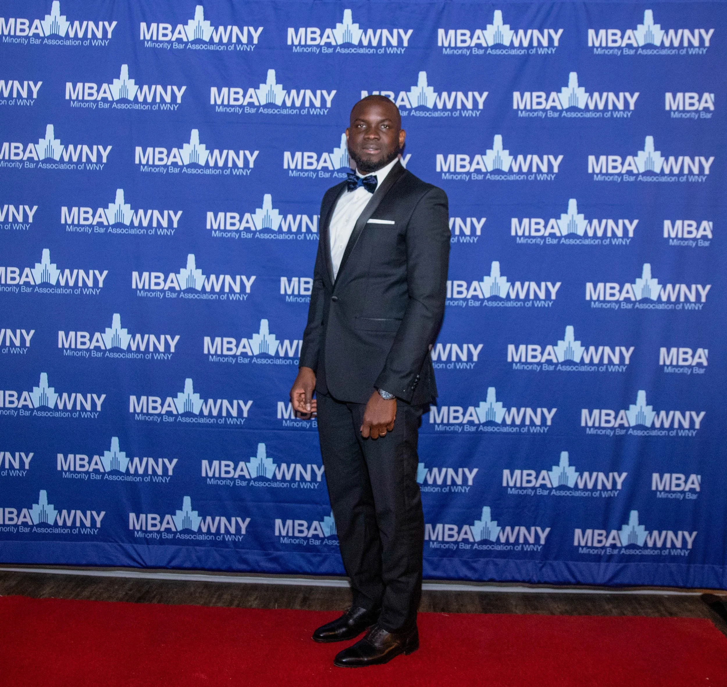 A man in a black tuxedo with a bowtie and dress shoes standing on a red carpet in front of a blue backdrop with the text "MBA WNY Minority Bar Association of WNY" repeated in a pattern.