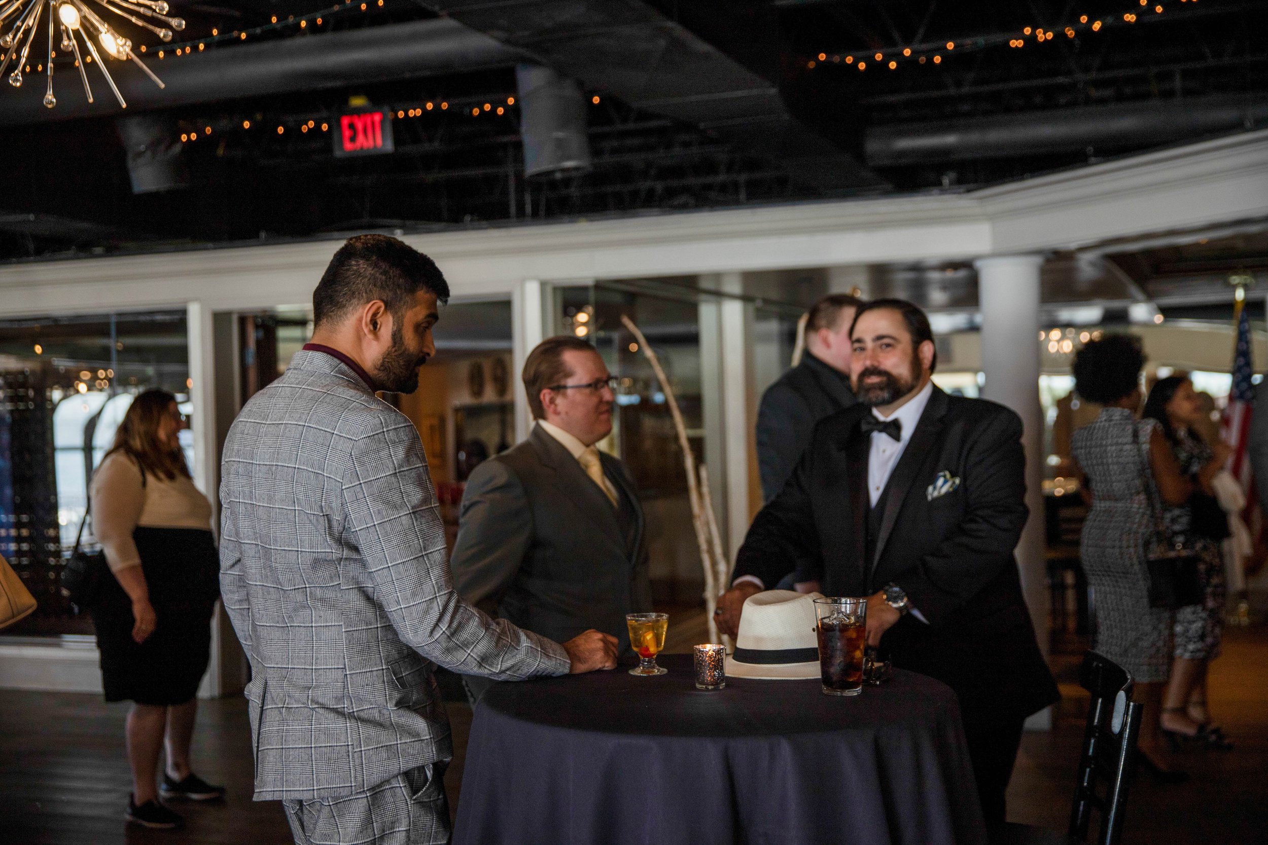 People socializing at a formal event with a bartender preparing drinks in the foreground, indoor setting with string lights and festive decorations.