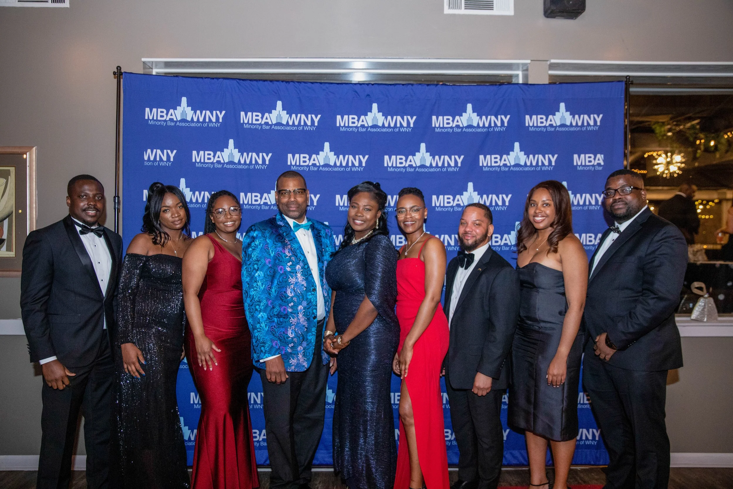 A group of nine individuals dressed in formal attire posing in front of a blue backdrop with the logo and text 'MBA WNY Minority Bar Association of WNY' for a photo at a formal event.