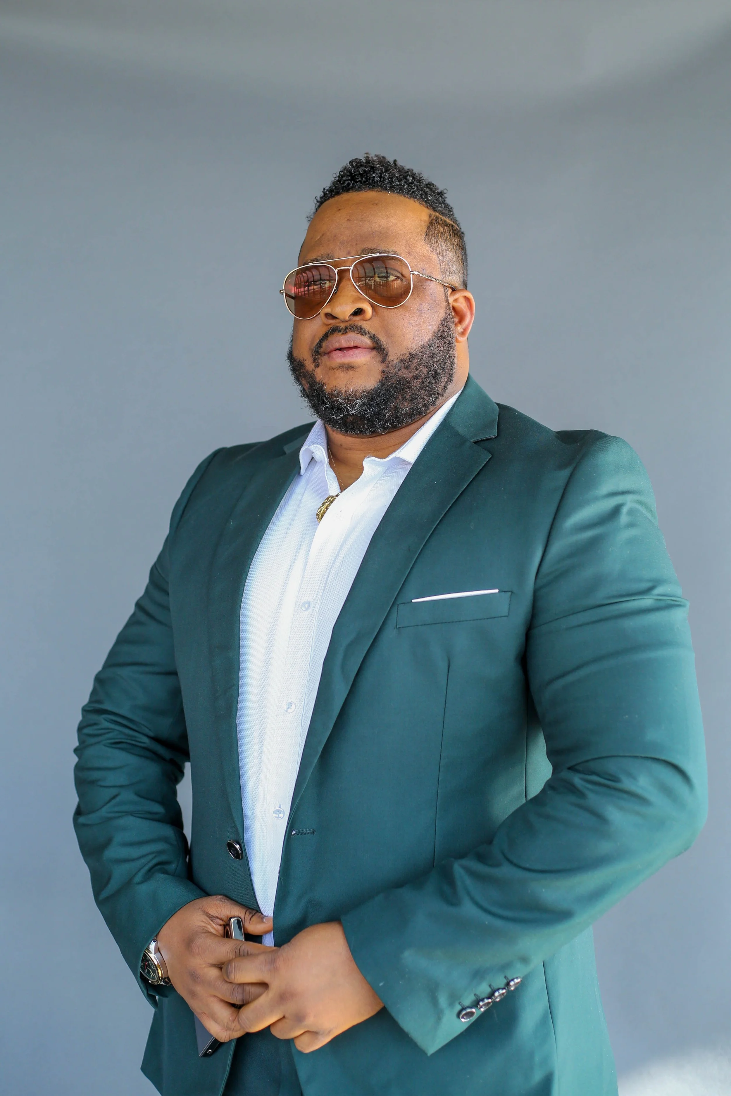 A man wearing sunglasses, a white shirt, and a teal suit jacket standing against a plain background.