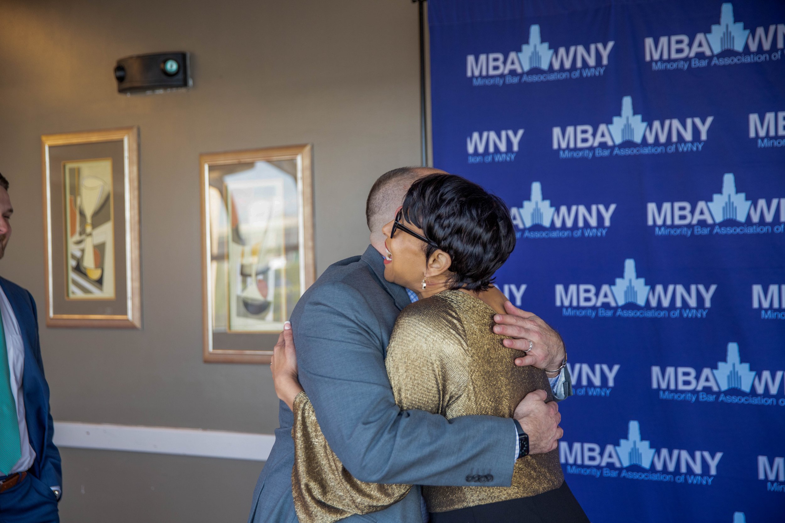 Two people hugging at a Minority Bar Association of WNY event, with a blue backdrop displaying the organization's logo.