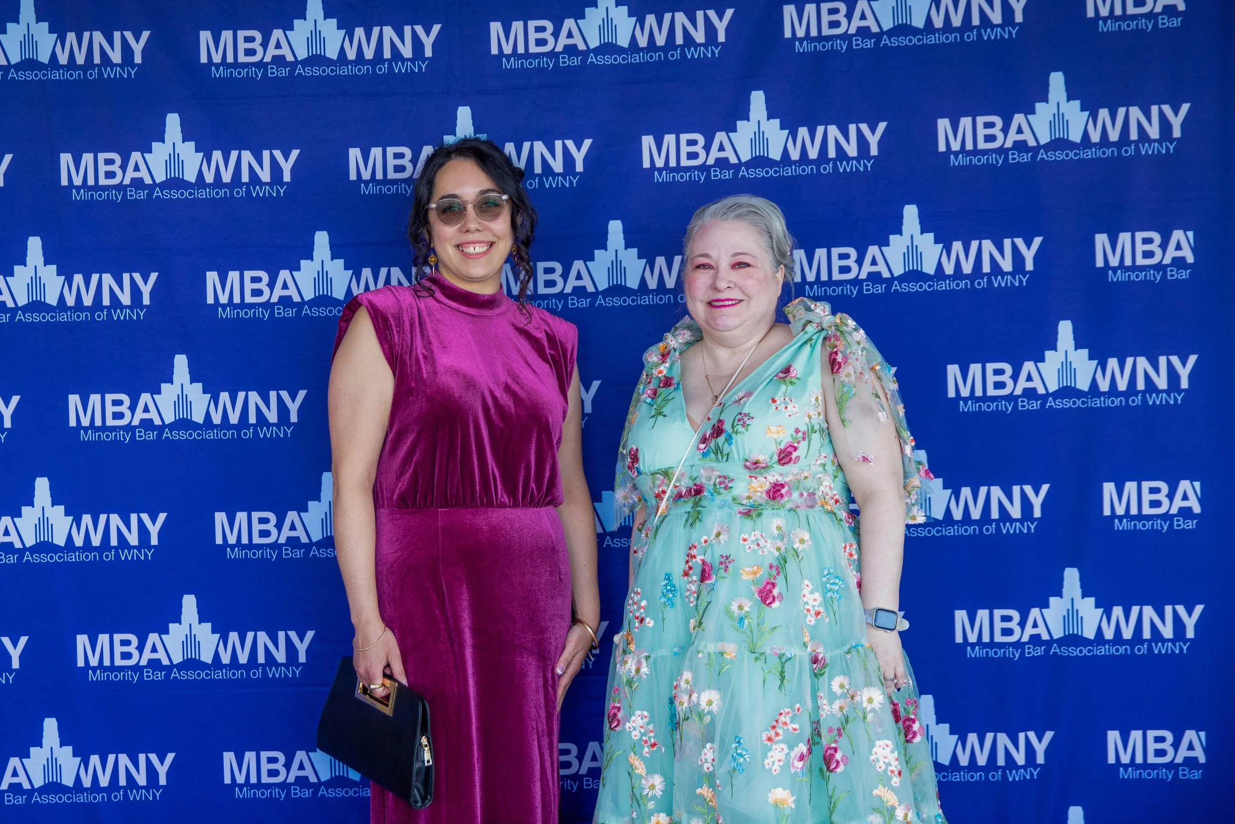 Two women standing in front of a blue backdrop with 'MBA WNY Minority Bar Association of WNY' logos. One woman is wearing a purple velvet dress with sunglasses, and the other woman is wearing a light blue floral dress with grey hair.