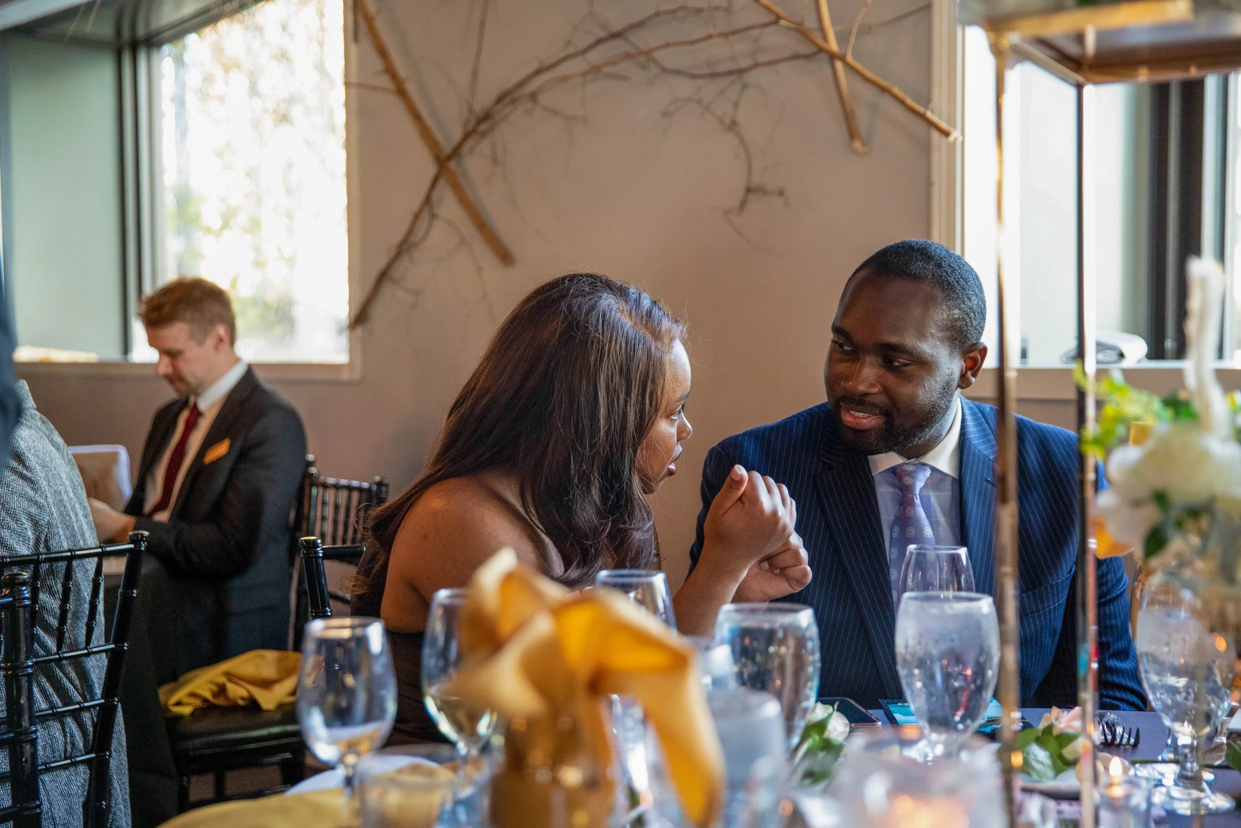 A group of people sitting at a dining table during a meal in a well-lit restaurant with large windows. One woman and one man are engaged in conversation, with glasses and tableware visible. There is a decorative branch on the wall behind them. The fo