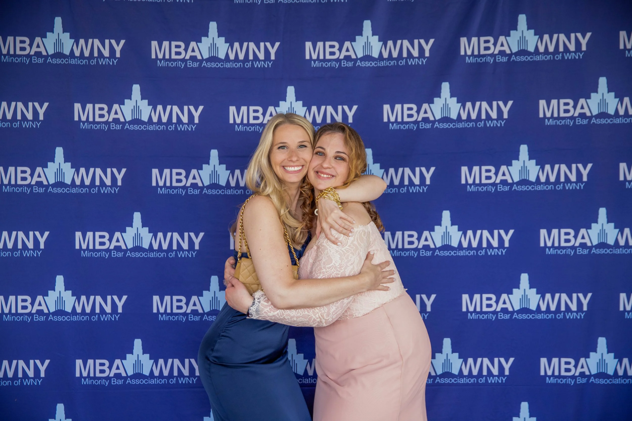 Two women hugging and smiling at a formal event with a blue backdrop that has the logo and text for the Minority Bar Association of WNY. One woman has blonde hair and is wearing a navy dress, the other has red hair and is wearing a pale pink dress.