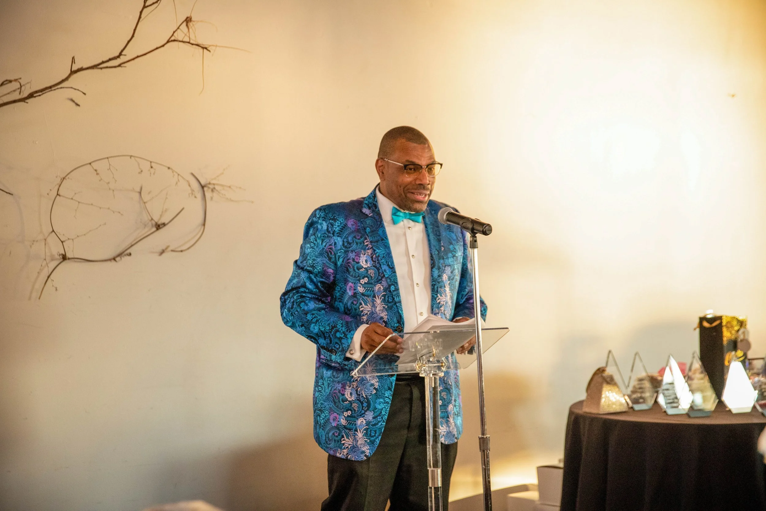 A man in a colorful blue patterned blazer with a blue bow tie speaking at a podium in a room with a plain light-colored wall. There are decorations and awards on a table nearby.