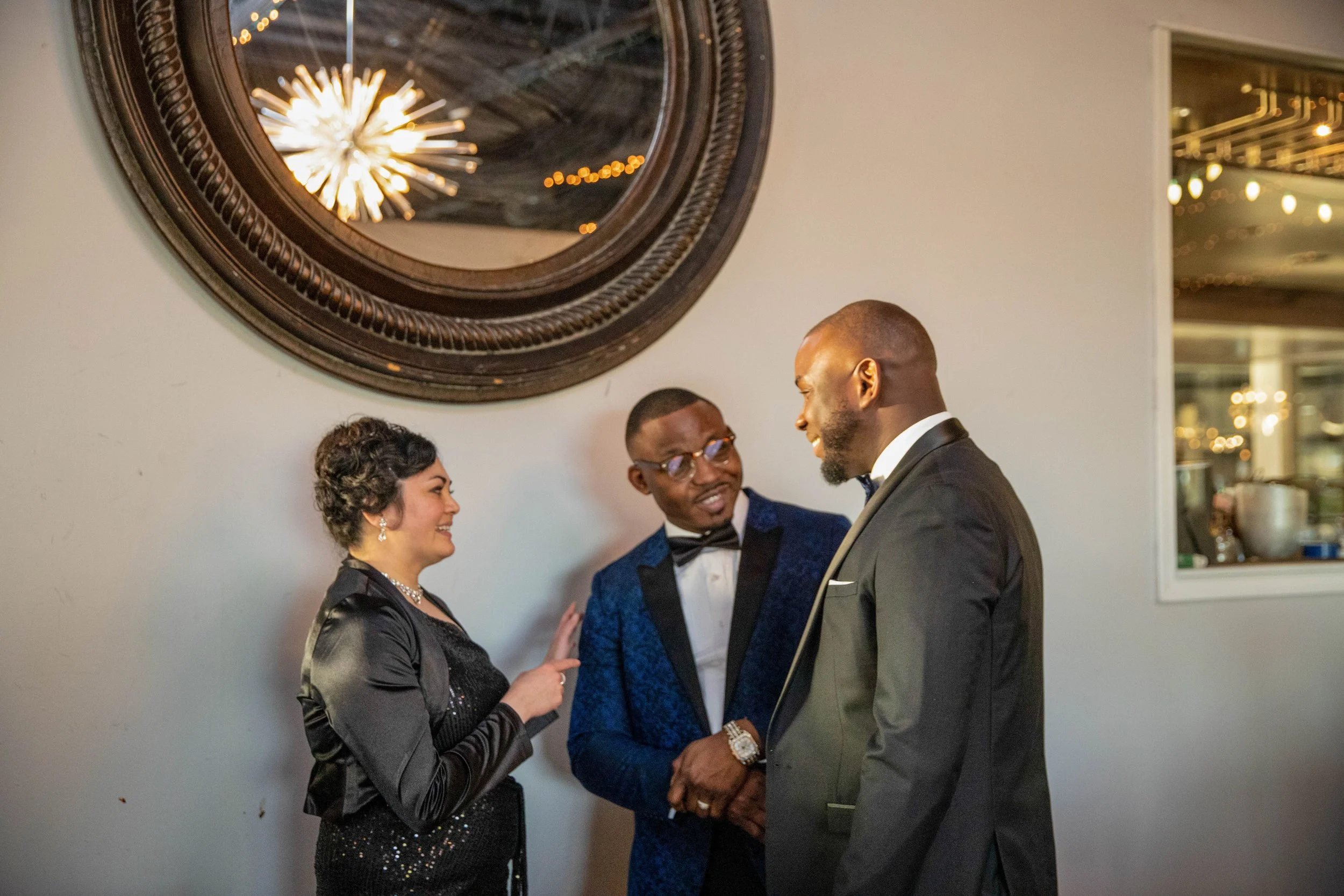 A group of three people, two men and one woman, dressed in formal attire, smiling, and engaging in conversation at a social event indoors. A large wall clock is visible above them.