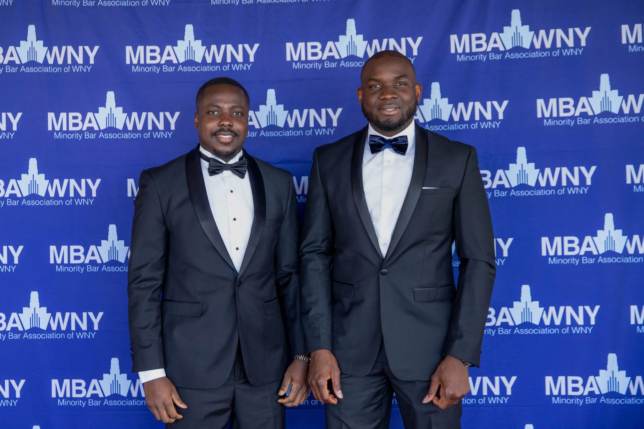 Two men in formal suits and bow ties standing in front of a blue backdrop with the logo and text of the Minority Bar Association of WNY.