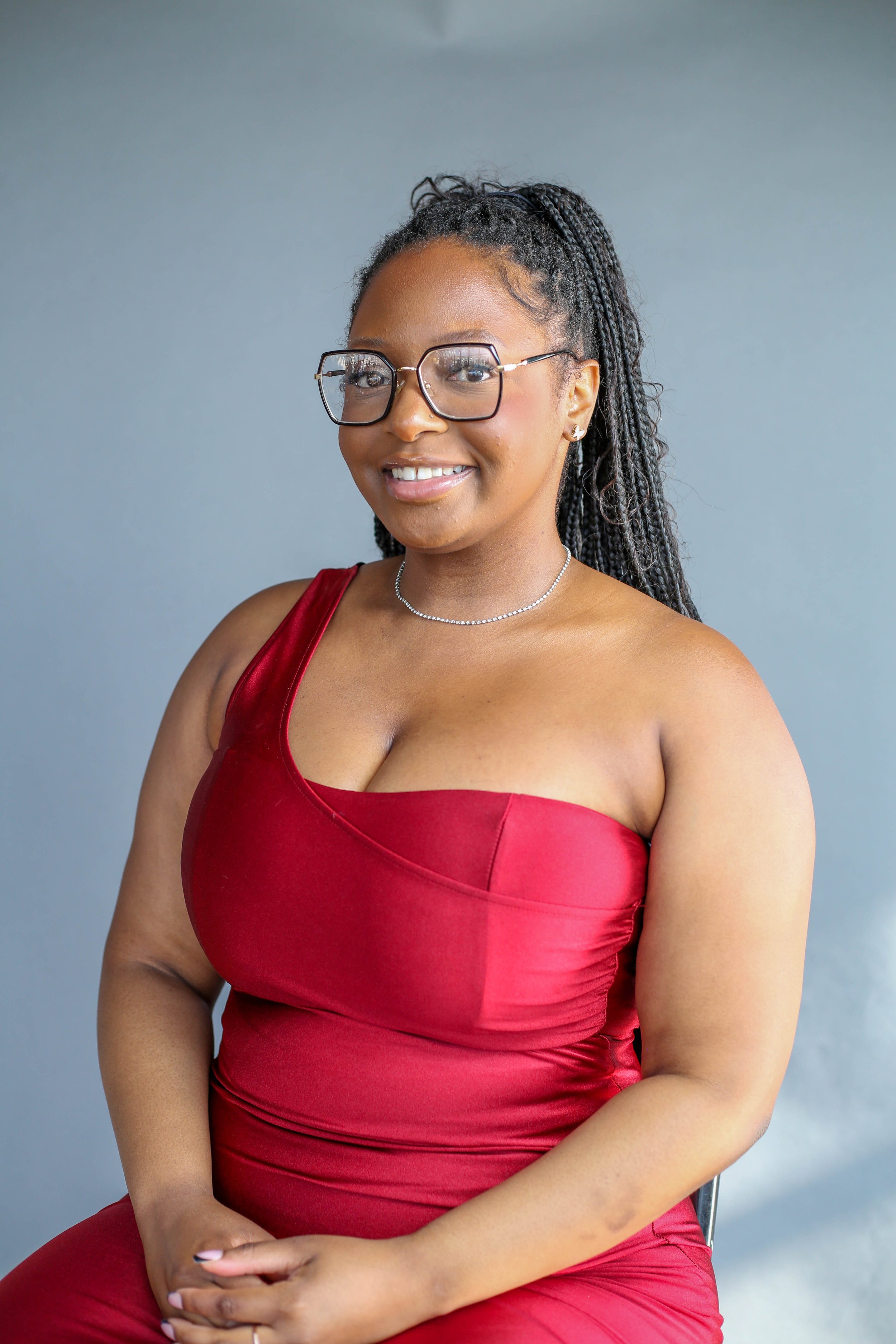 A smiling woman with glasses and braided hair wearing a red dress and silver necklace, posing against a plain background.