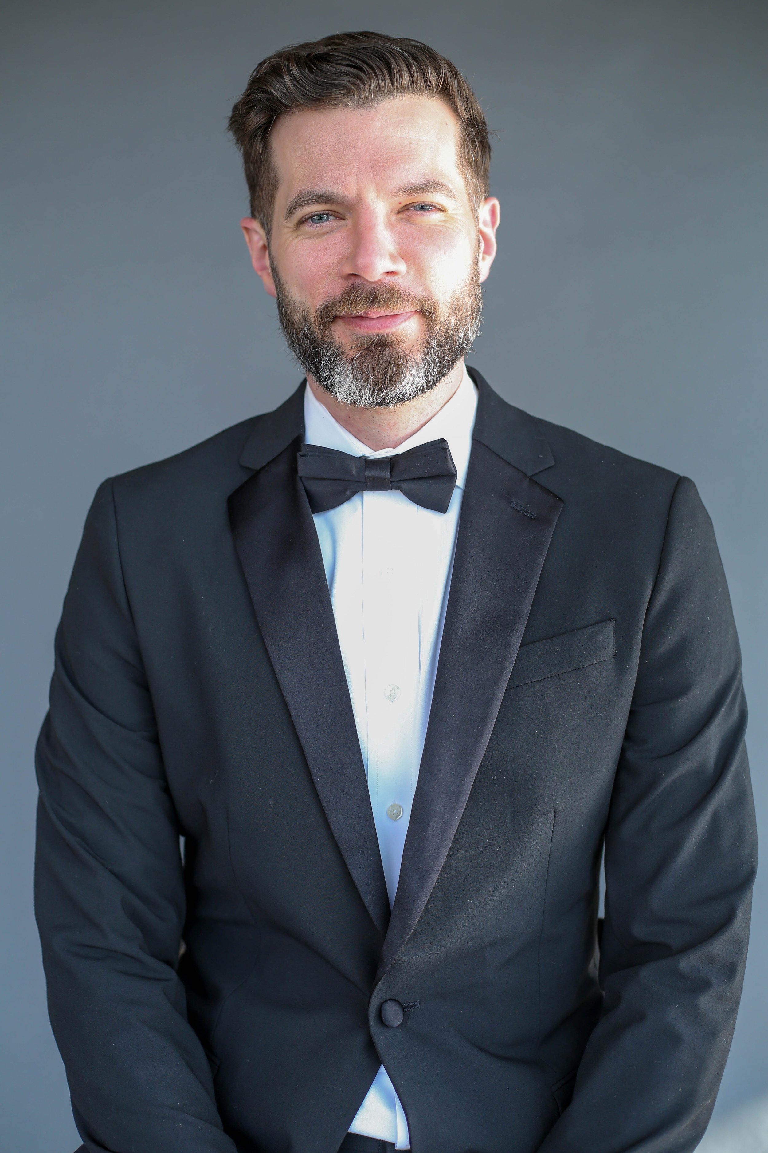 A man with a beard and short hair, dressed in a black tuxedo with a bow tie, smiling at the camera against a gray background.
