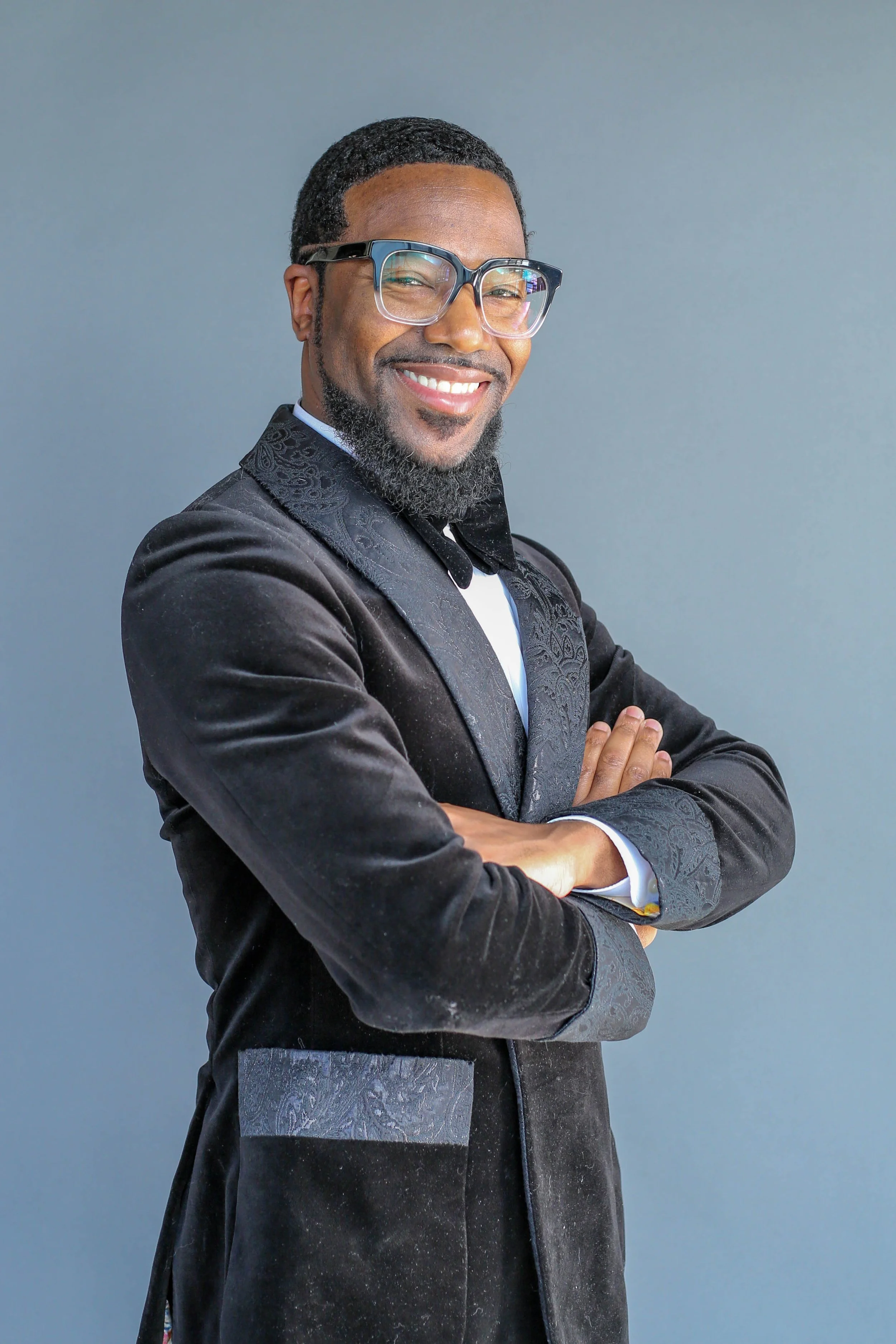 A smiling man with glasses and a beard, wearing a black suit jacket with embroidered details and gray pants, standing with crossed arms against a plain gray background.