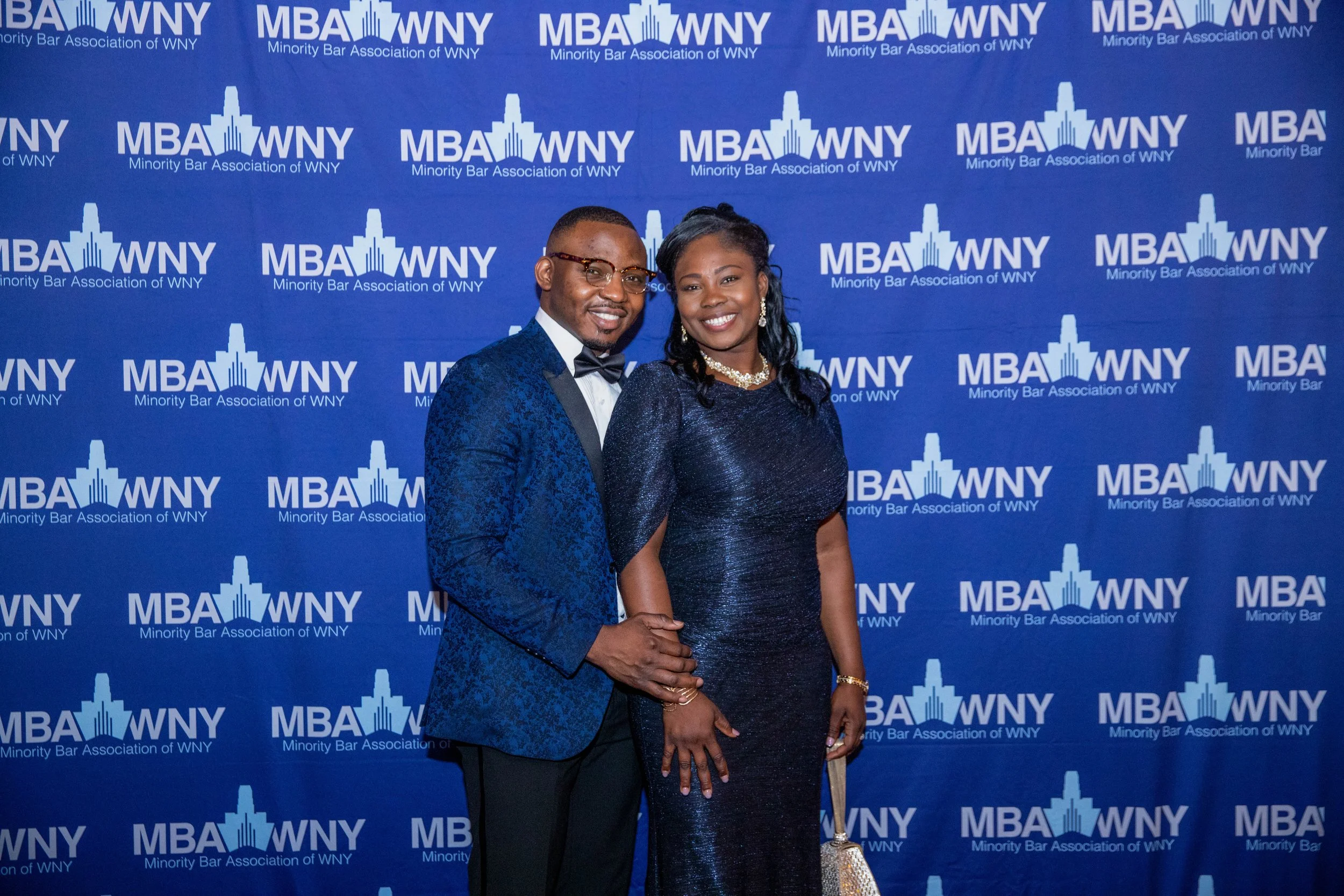 A man and a woman dressed in formal attire standing together and smiling at an event with a blue backdrop that has the logos of the Minority Bar Association of WNY and the MBA WNY repeated across it.