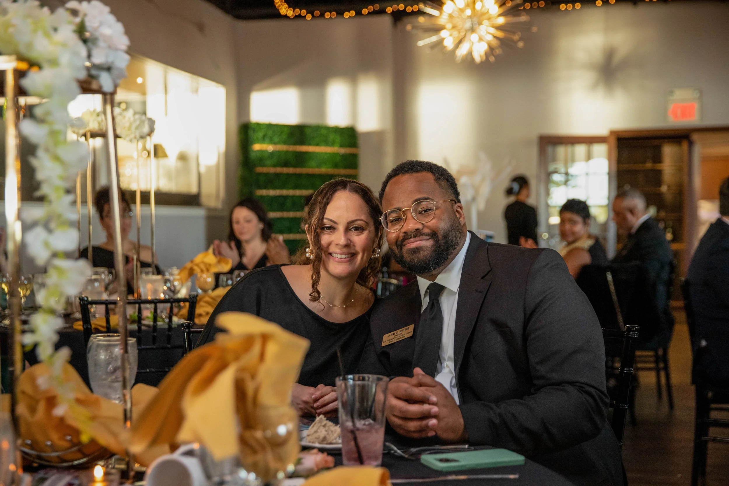 Smiling couple dressed in formal attire at a decorated event, sitting at a table with food and drinks, surrounded by other guests in a well-lit venue with floral decorations.
