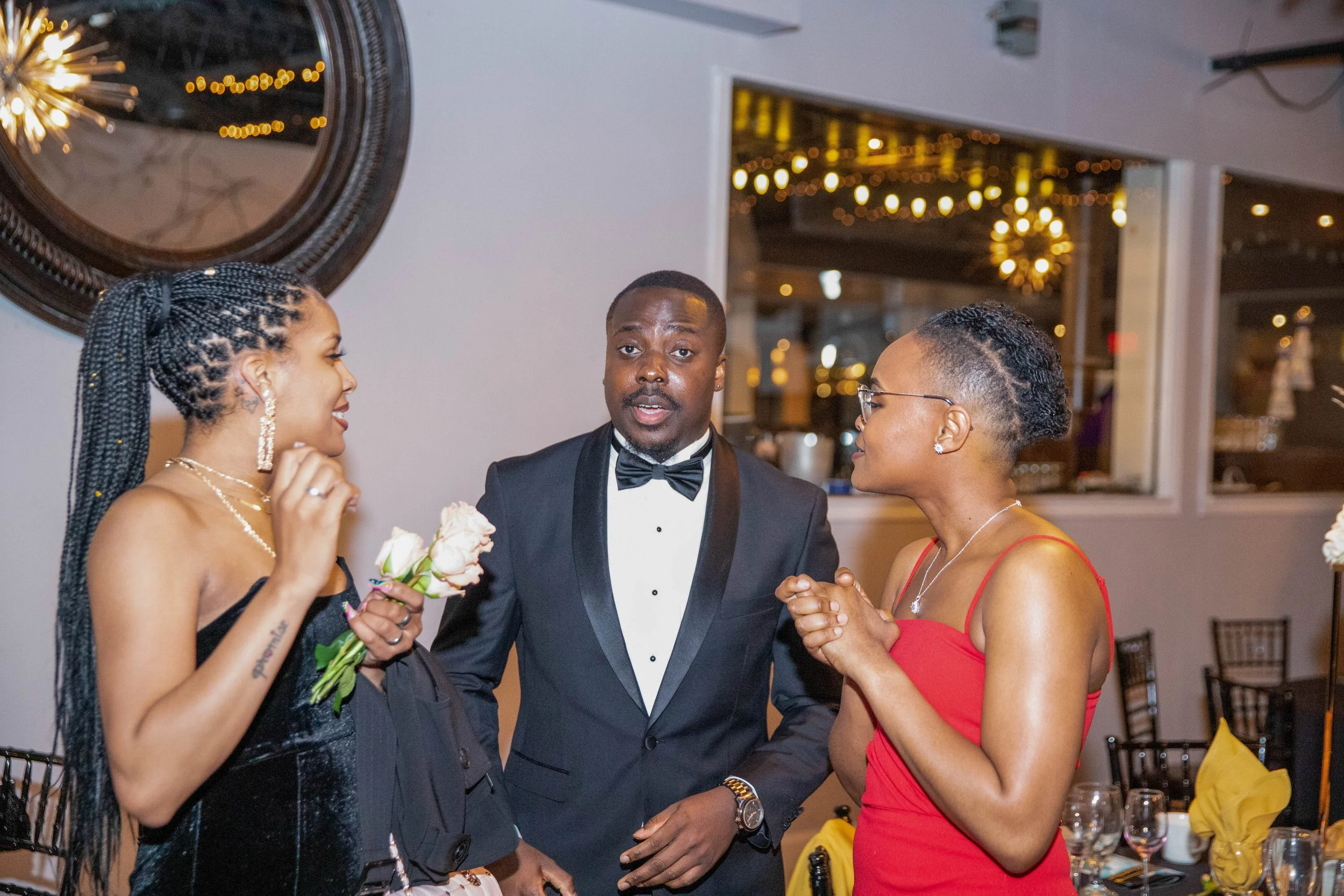 Three women and a man dressed in formal attire talking and smiling at an indoor event with decorative lighting and tables with yellow napkins in the background.