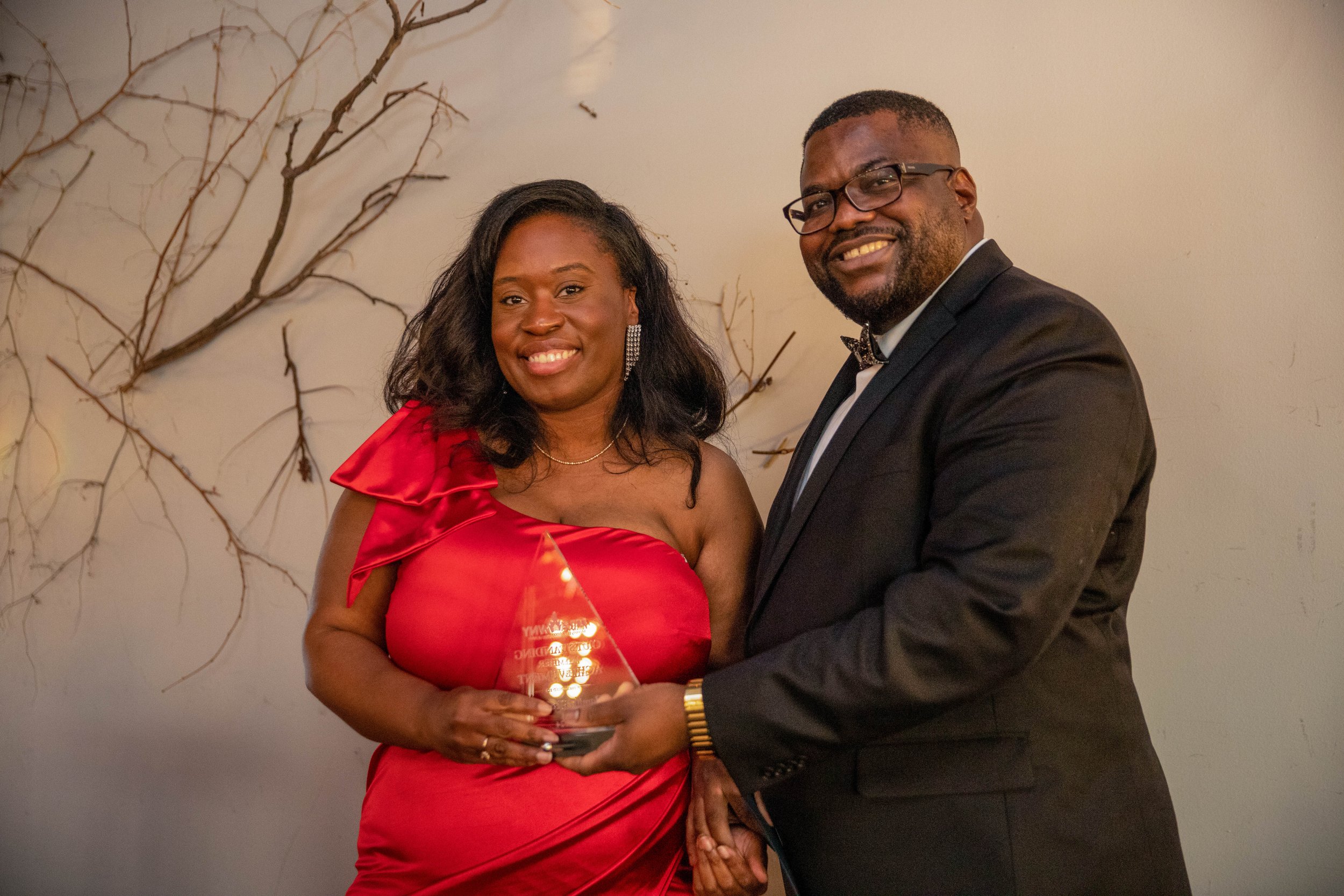 A smiling woman in a red dress holds a glass award, standing next to a smiling man in a black tuxedo with a bow tie and glasses. They are both facing the camera, and the background features a wall with decorative, leafless tree branches.