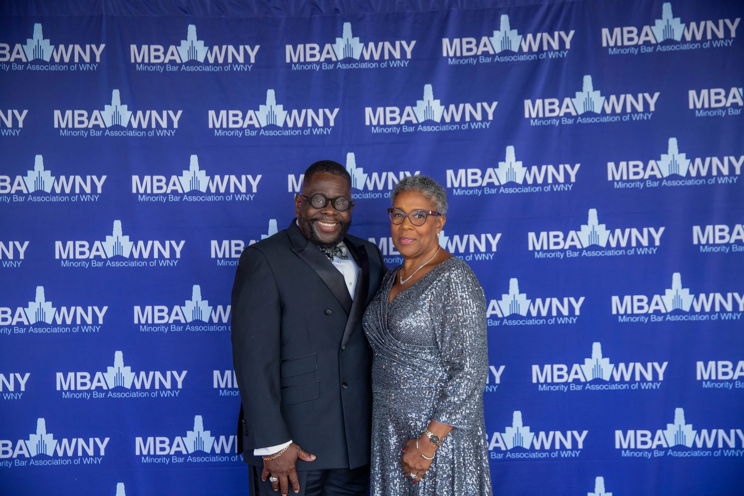 Two individuals standing together at an event with a blue backdrop that has the logo and name of the Minorit Bar Association of WNY repeated across it. The man on the left is wearing a dark tuxedo and glasses, and the woman on the right is wearing a 