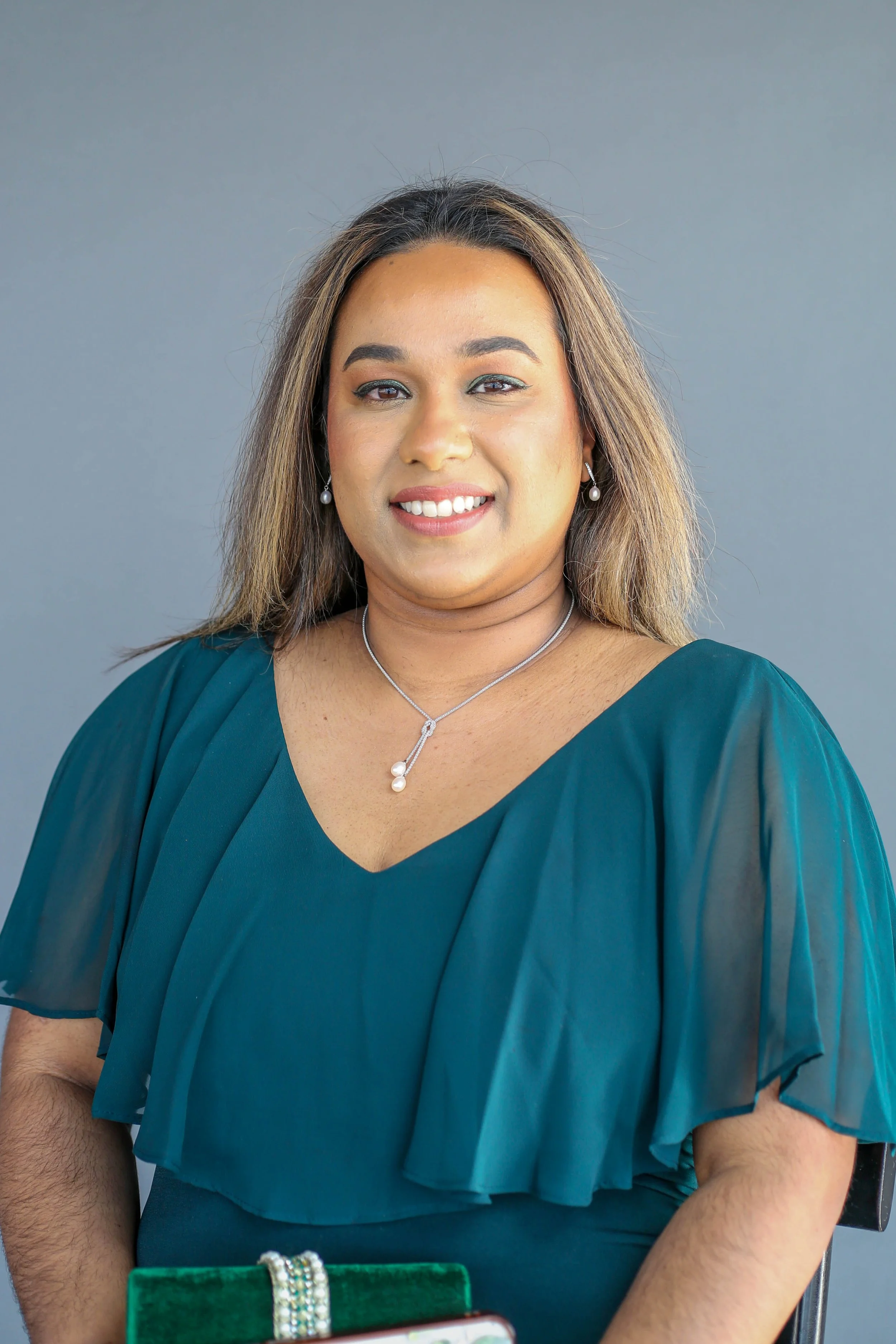 A woman with shoulder-length hair, wearing a teal dress, pearl earrings, and a silver necklace with a pearl pendant, smiling at the camera against a plain gray background.