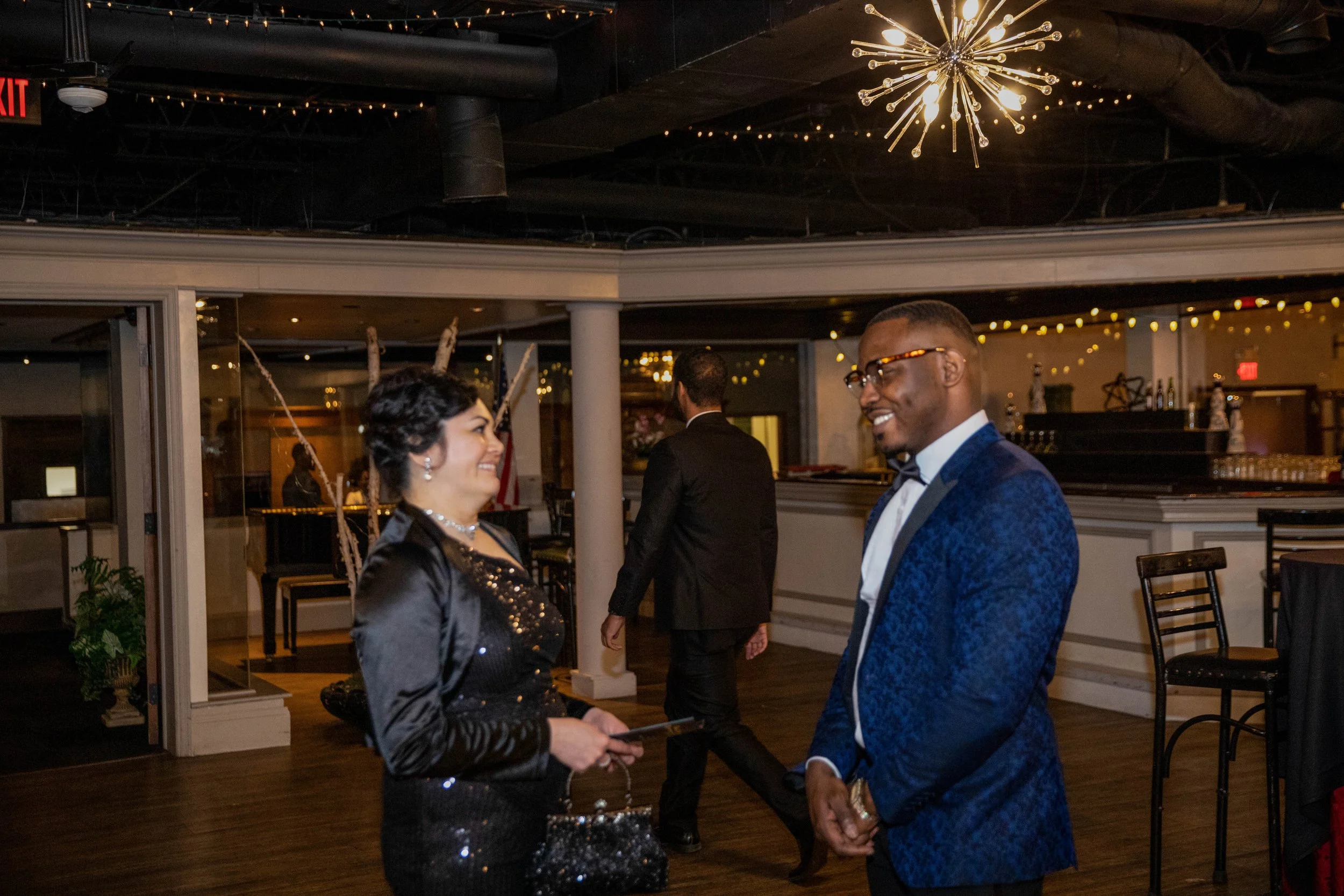 A man and woman are smiling and talking at a formal event in a dimly lit room with decorative lighting and a modern chandelier.